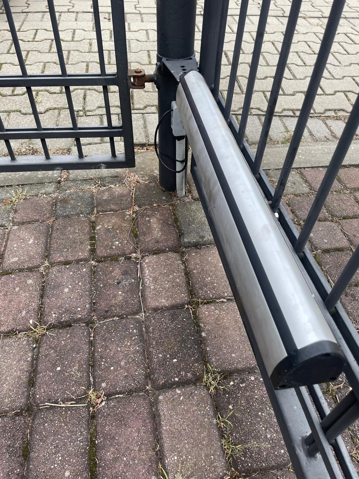 Automatyka otwierająca czarną, metalową bramę, widoczny siłownik z przewodami na tle kostki brukowej.