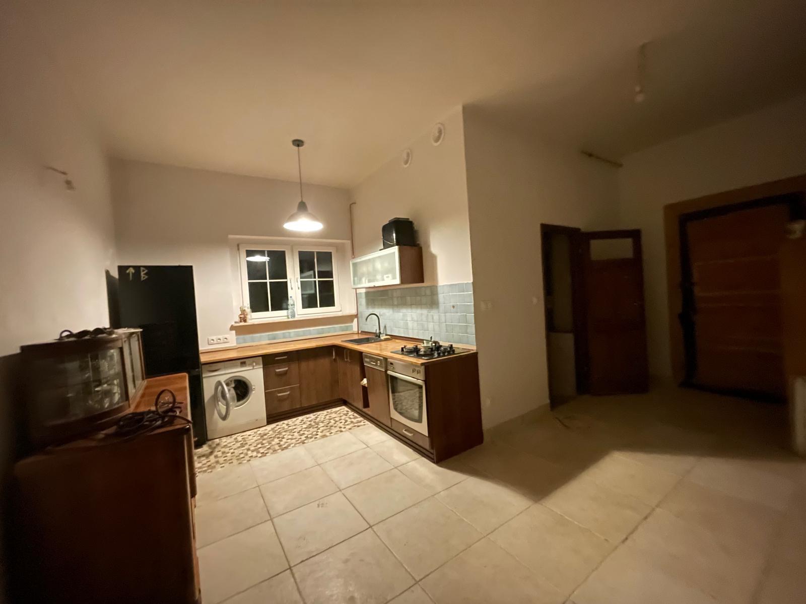 Ukończona aranżacja kuchni z drewnianymi szafkami, płytkami w odcieniach błękitu nad blatem, pralką w zabudowie i lampą wiszącą w stylu retro, widok na otwarty salon z drzwiami.