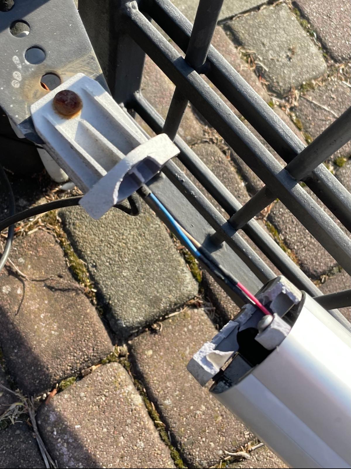 Uszkodzony mechanizm automatycznej bramy wjazdowej z widocznymi przewodami, mocowany do metalowego ogrodzenia na tle kostki brukowej.