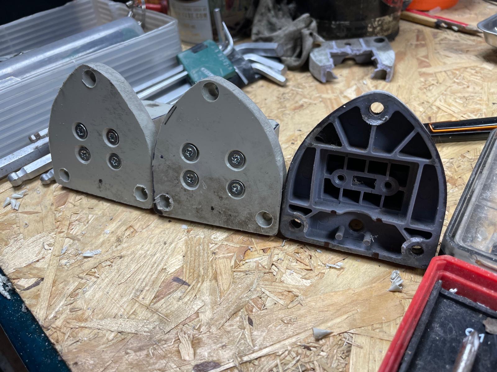 Trzy uchwyty montażowe, dwa szare z wkrętami i jeden szary z widoczną konstrukcją wewnętrzną, leżące na drewnianym blacie roboczym, otoczone narzędziami.