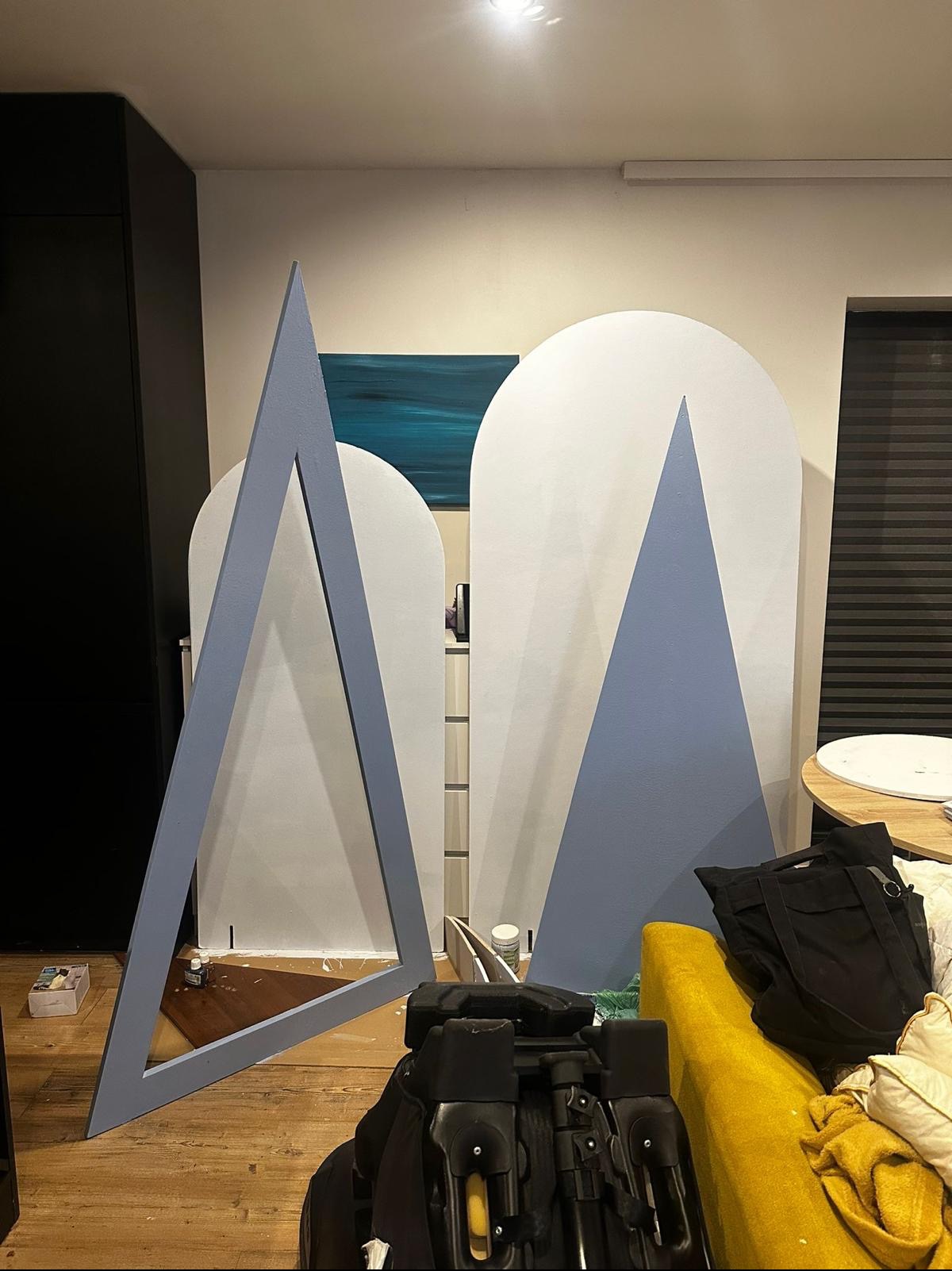 Przygotowane do sesji zdjęciowej, białe, łukowate tła z niebieskimi trójkątami, ustawione w pomieszczeniu z drewnianą podłogą, widoczne rekwizyty i torba. W tle obraz z motywem morskim.