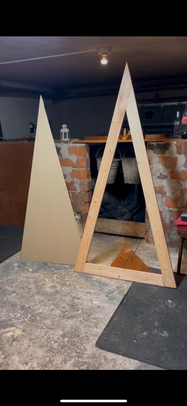 Dwa trójkąty: jeden z płyty, drugi z surowego drewna, ustawione w pomieszczeniu z betonową podłogą i ścianą z cegieł w tle, prawdopodobnie w trakcie tworzenia dekoracji lub elementu konstrukcyjnego.