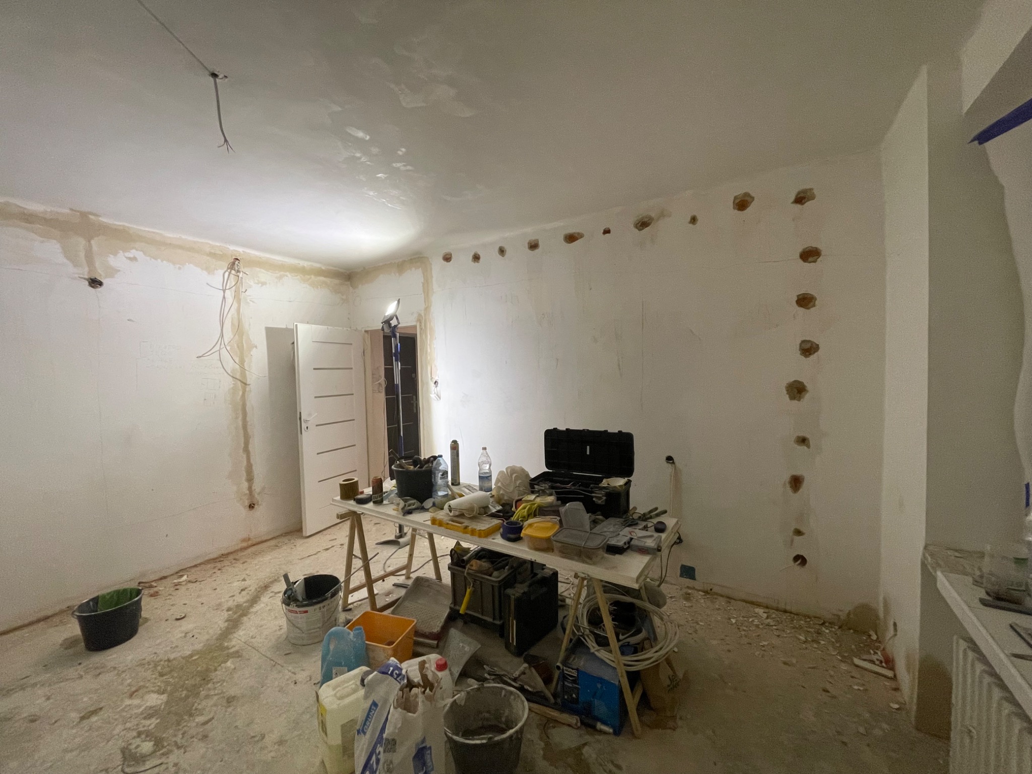 Pokój w trakcie prac przygotowawczych do malowania: ściany z widocznymi śladami po szpachlowaniu i wierceniu, prowizoryczny stół roboczy zastawiony narzędziami, wiadra i pojemniki z materiałami...