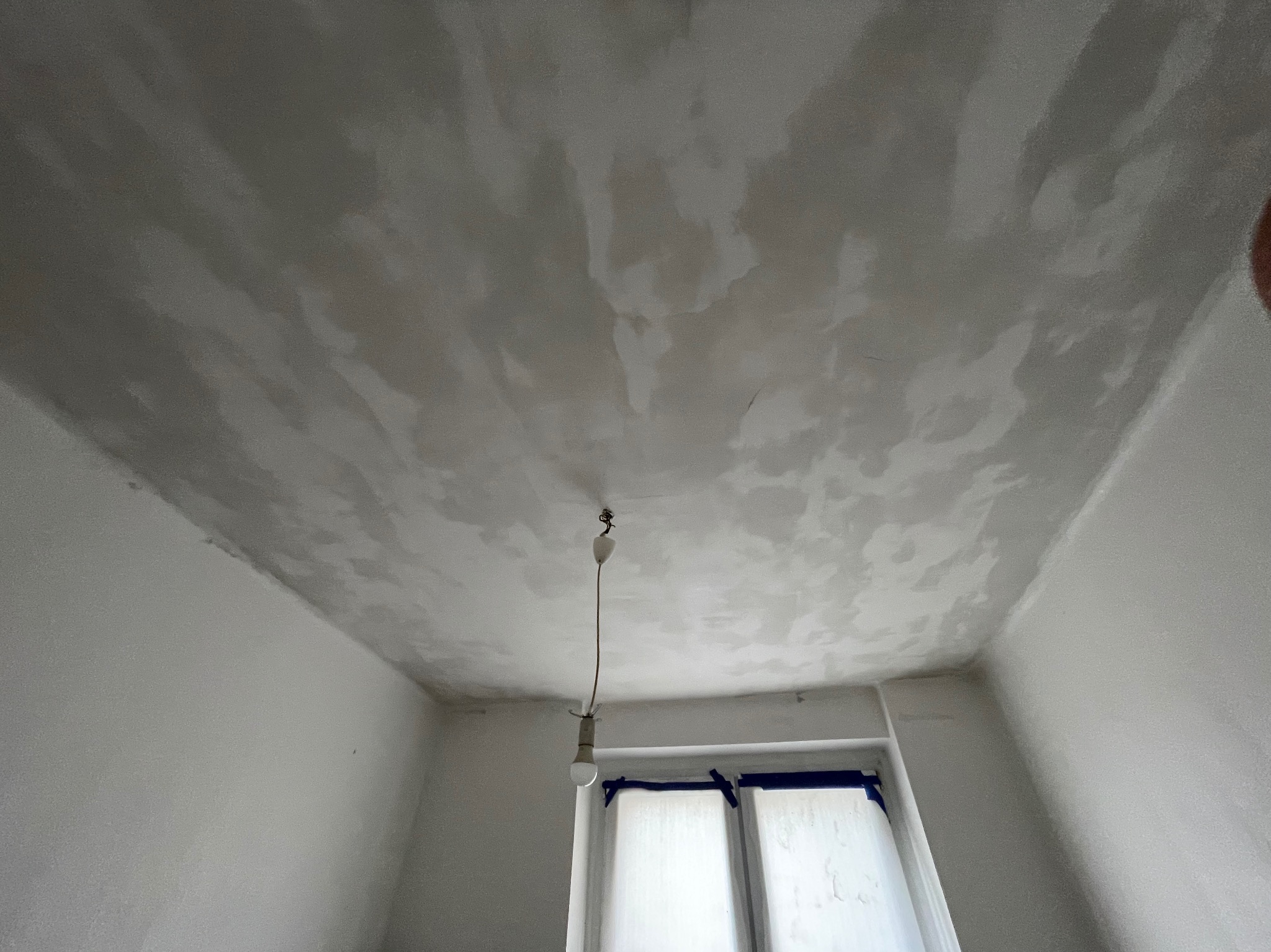 Szpachlowana sufit i ściany w pokoju, przygotowane do malowania, widoczne okno z zabezpieczeniem folią i taśmą malarską oraz wisząca żarówka.