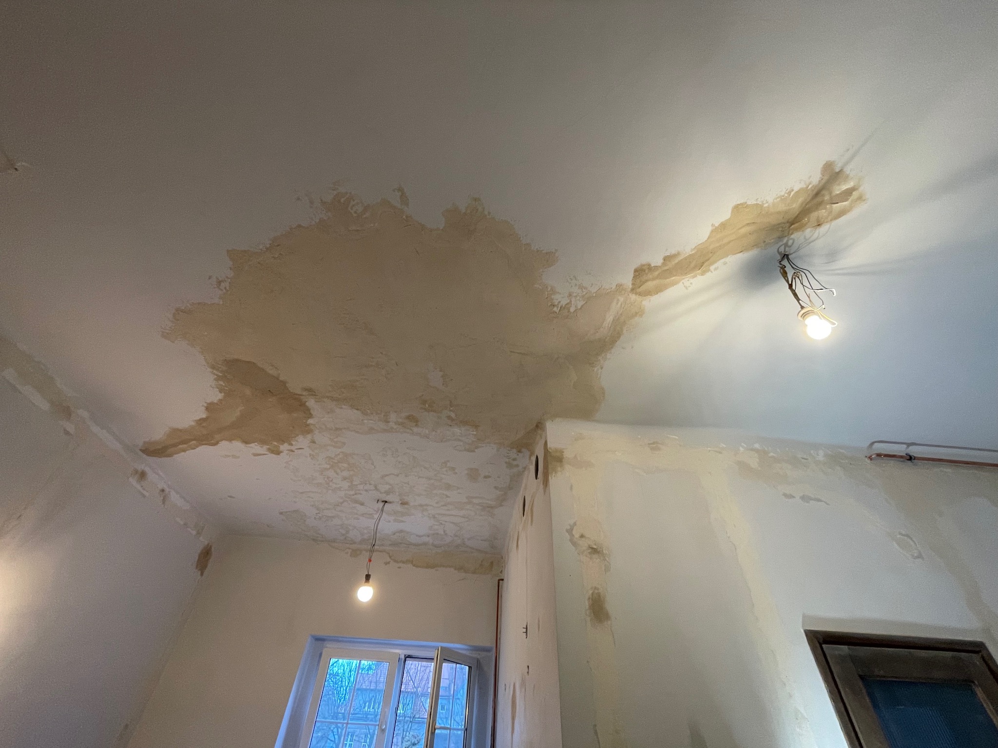 Zacieki na suficie i ścianach w rogu pomieszczenia, widoczne ślady wilgoci i odpadająca farba, odsłonięte przewody z żarówką.