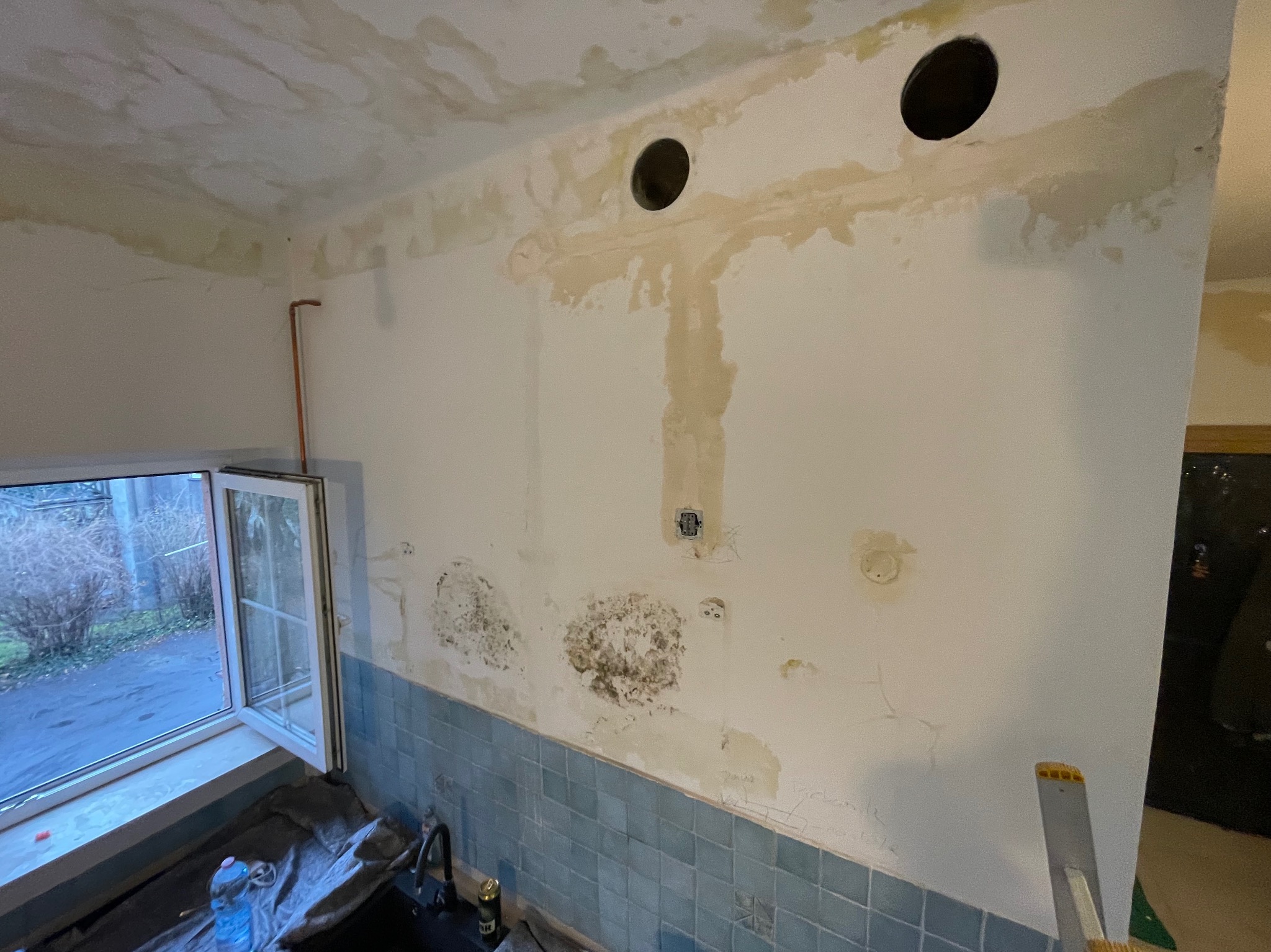 Ściana w łazience przed remontem z widocznymi śladami wilgoci, grzybem i przygotowanymi otworami wentylacyjnymi, niebieskie kafelki na dole ściany, okno z widokiem na zewnątrz.