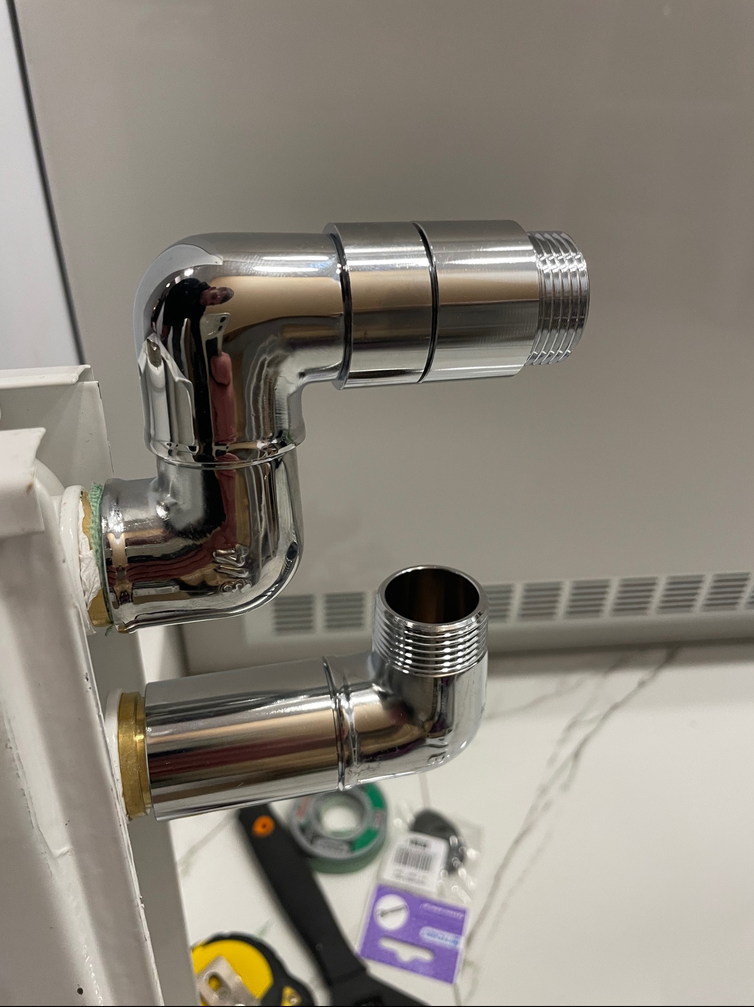 Błyszczące chromowane rury łączące grzejnik, widoczna taśma uszczelniająca i narzędzia na marmurowym blacie.