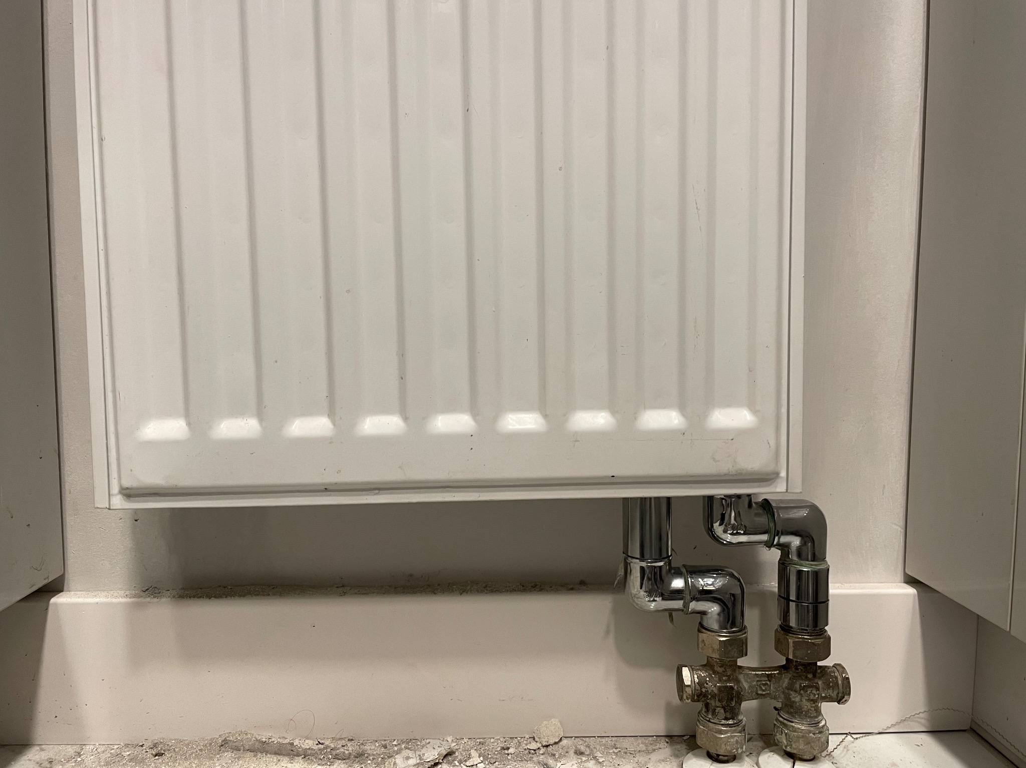 Biały grzejnik panelowy z chromowanymi rurami i zaworami przy podłodze w trakcie prac remontowych, widoczne zabrudzenia i resztki materiałów budowlanych.