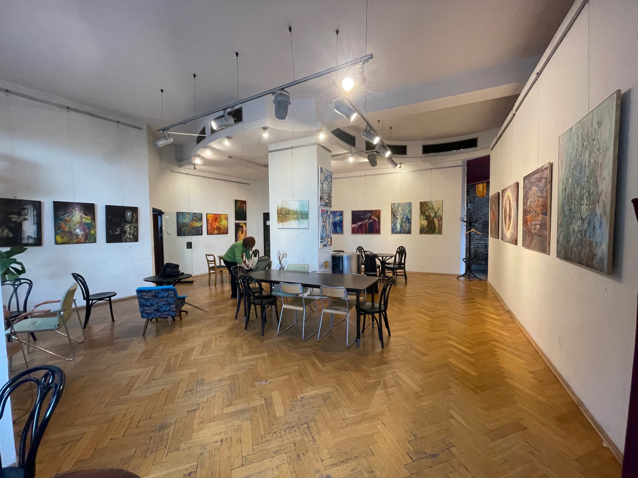 Przestronne wnętrze galerii sztuki z parkietem w jodełkę, oświetleniem punktowym i rzędami obrazów na ścianach, gdzie osoba w zielonej bluzce ustawia wazony na stole.