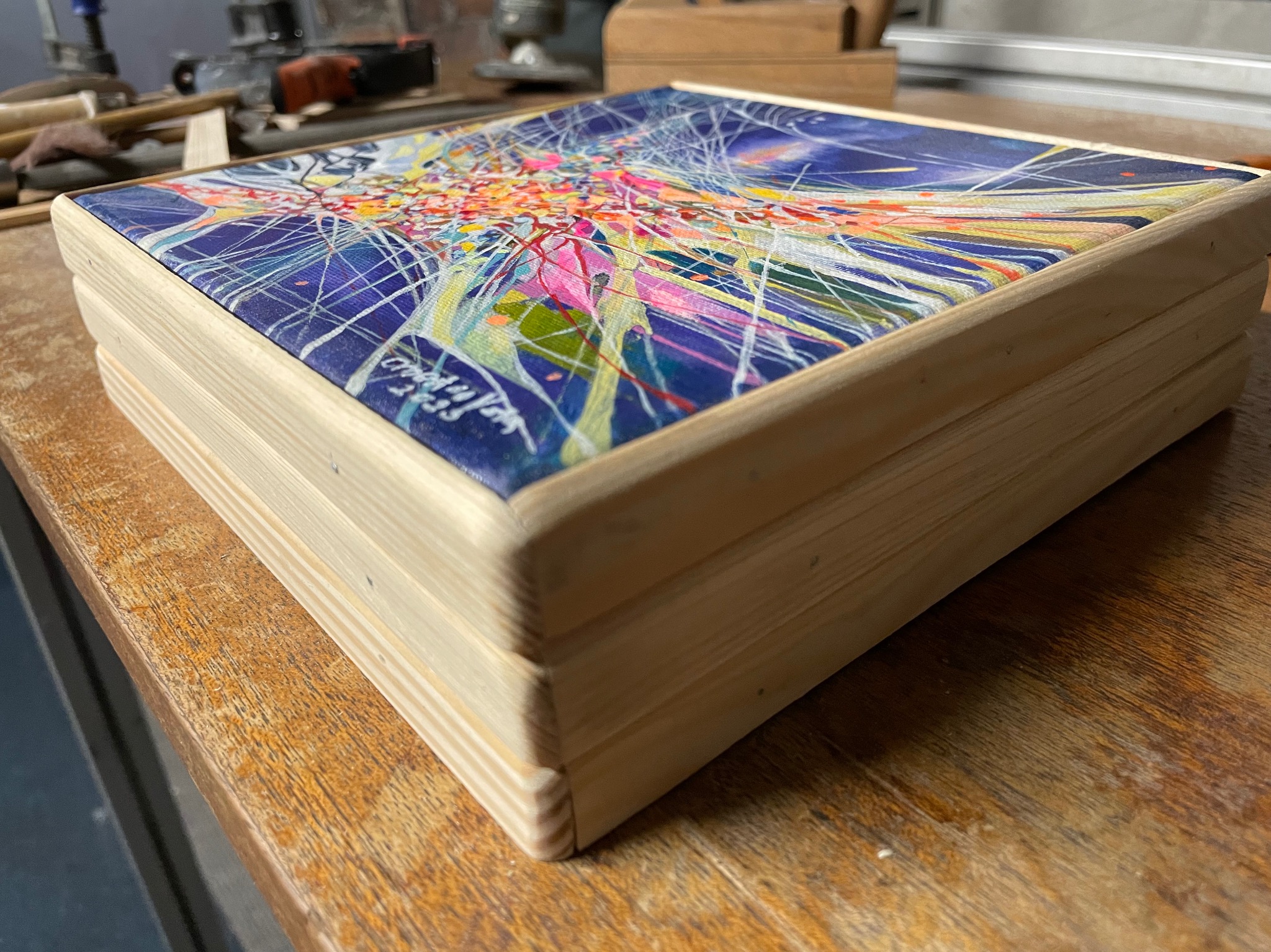 Drewniana szkatułka z abstrakcyjnym, kolorowym malunkiem na wieczku, leżąca na drewnianym blacie roboczym. Widoczne ślady użytkowania na blacie i niedoskonałości w wykończeniu szkatułki.