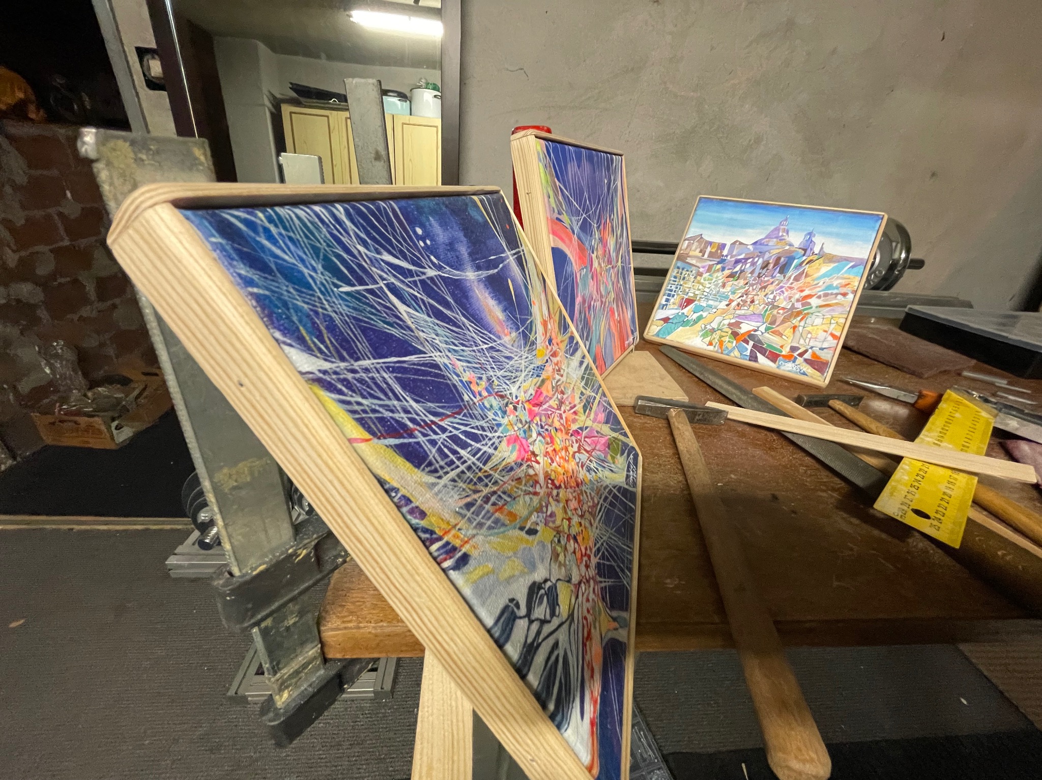 Trzy obrazy na drewnianych blejtramach, jeden w imadle, na stole z narzędziami, w warsztacie.