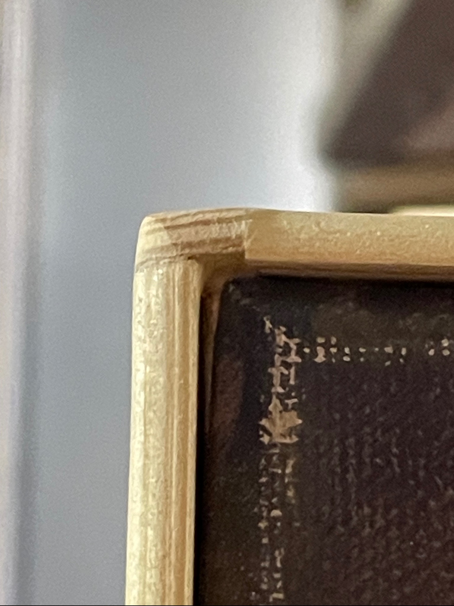 Fragment rogu drewnianej ramy obrazu z widocznymi śladami zużycia i odpryskami farby, na tle jasnej ściany.