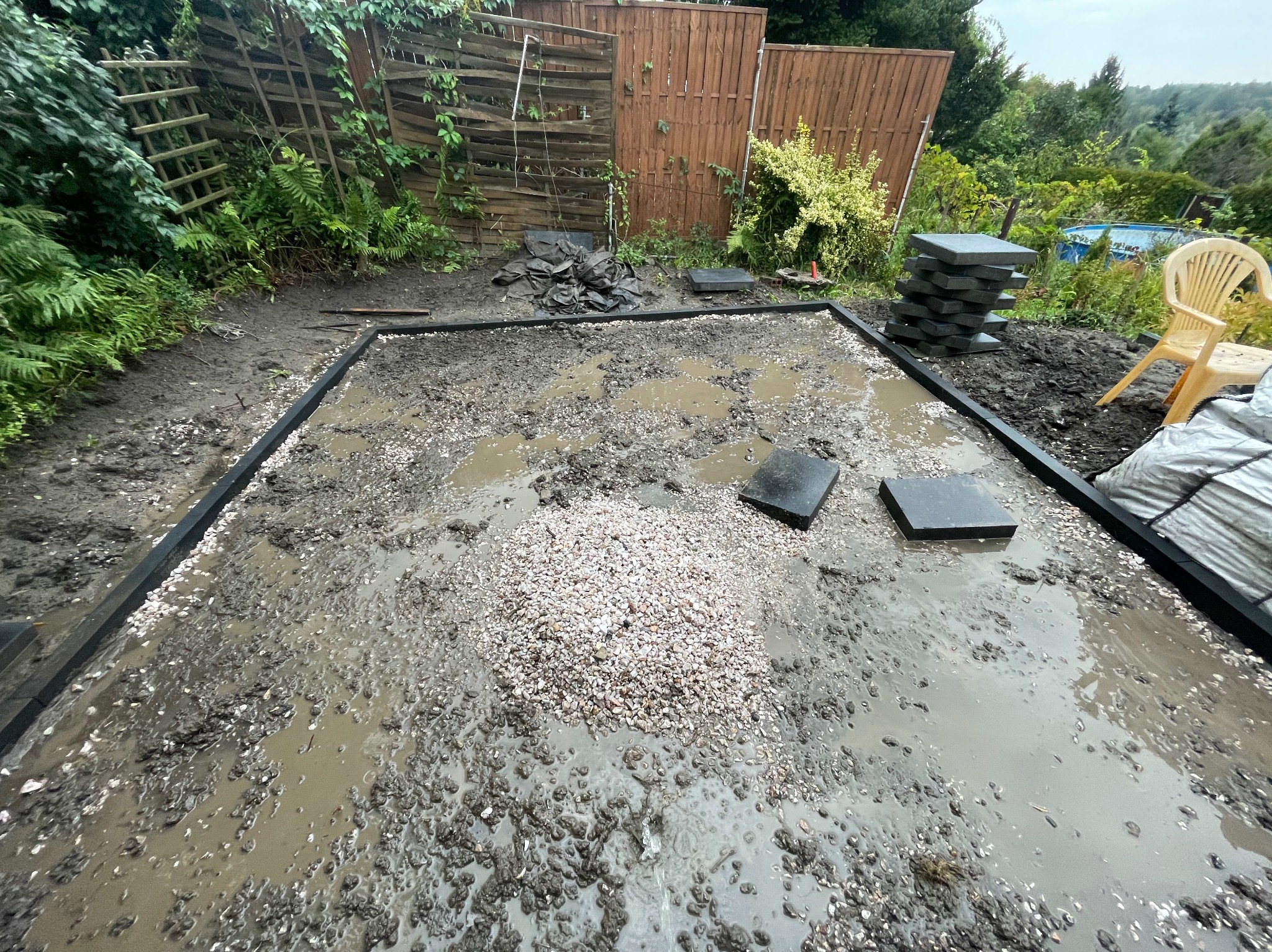 Przygotowanie terenu pod taras lub ścieżkę: wytyczony obrys z czarnych obrzeży, wysypany żwir, ułożone płyty chodnikowe, widoczna sterta płyt i krzesło ogrodowe w tle.