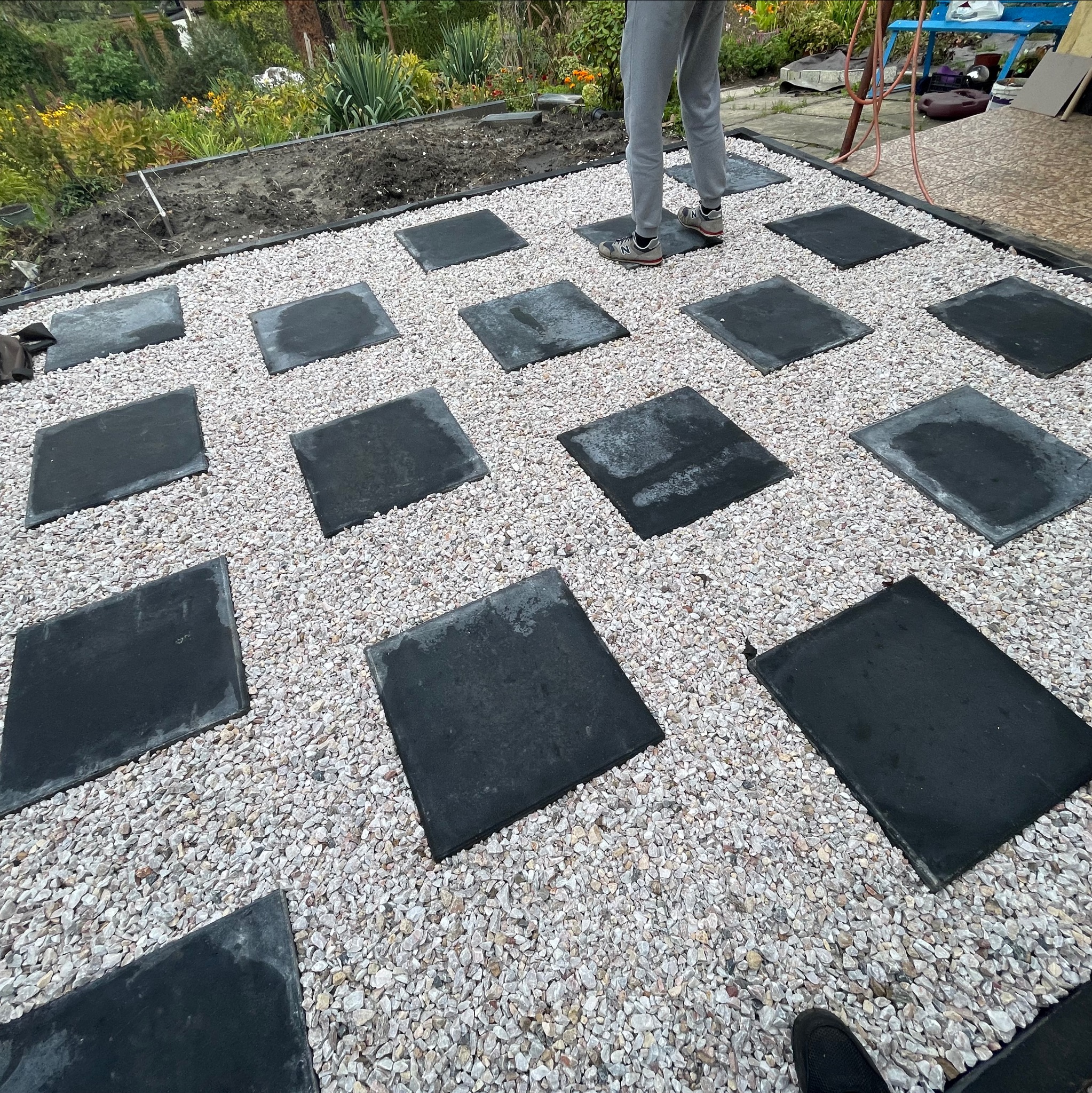 Układanie ścieżki z kwadratowych płyt na jasnym żwirze w ogrodzie, widoczne fragmenty roślinności w tle, osoba w szarych spodniach w trakcie pracy.