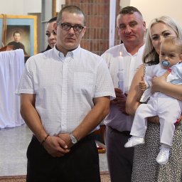 Igłą i Ołówkiem MONIKA LAMPERT - Rodzina z dzieckiem podczas uroczystości w kościele, w tle obraz religijny i świece. Mama trzyma niemowlę ubrane w biały strój.