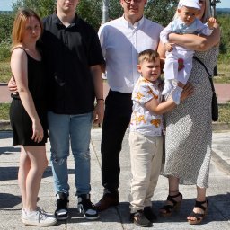 Igłą i Ołówkiem MONIKA LAMPERT - Rodzinne zdjęcie grupowe na zewnątrz w słoneczny dzień. Dwie kobiety, dwóch mężczyzn i dwoje dzieci pozują do zdjęcia na tle zieleni i błękitnego nieba.