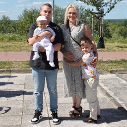 Igłą i Ołówkiem MONIKA LAMPERT - Rodzinne zdjęcie plenerowe: młody mężczyzna trzyma niemowlę w białym ubranku, obok kobieta w sukience i starszy chłopiec. W tle zieleń i rzeka.