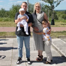 Rodzinne zdjęcie plenerowe: młody mężczyzna trzyma niemowlę w białym ubranku, obok kobieta w sukience i starszy chłopiec. W tle zieleń i rzeka.