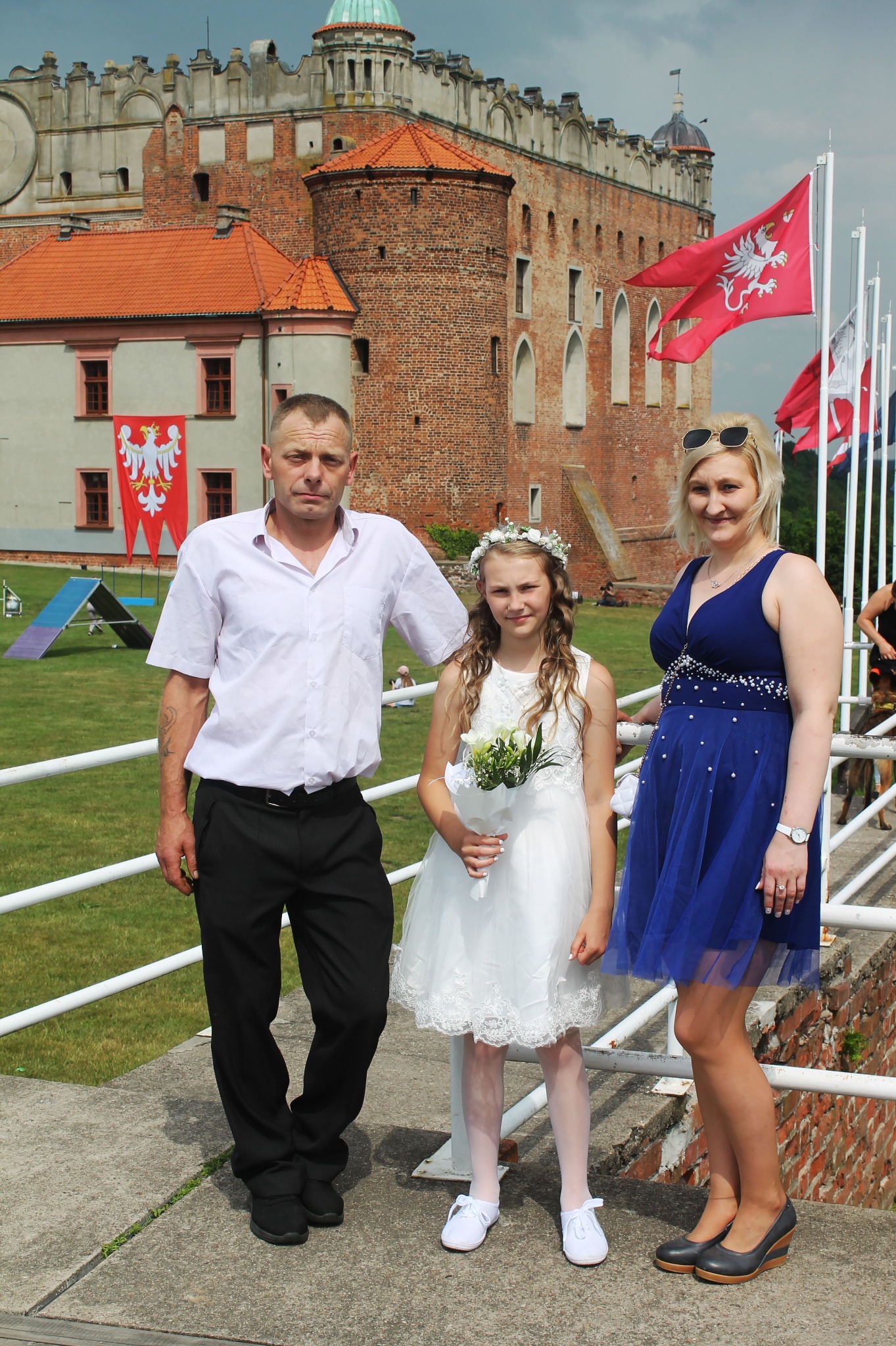 Rodzinne zdjęcie przed gotyckim zamkiem w Gniewie: dziewczynka w białej sukience z bukiecikiem, mężczyzna w koszuli i kobieta w niebieskiej sukience pozują na tle historycznej budowli i polskich flag.