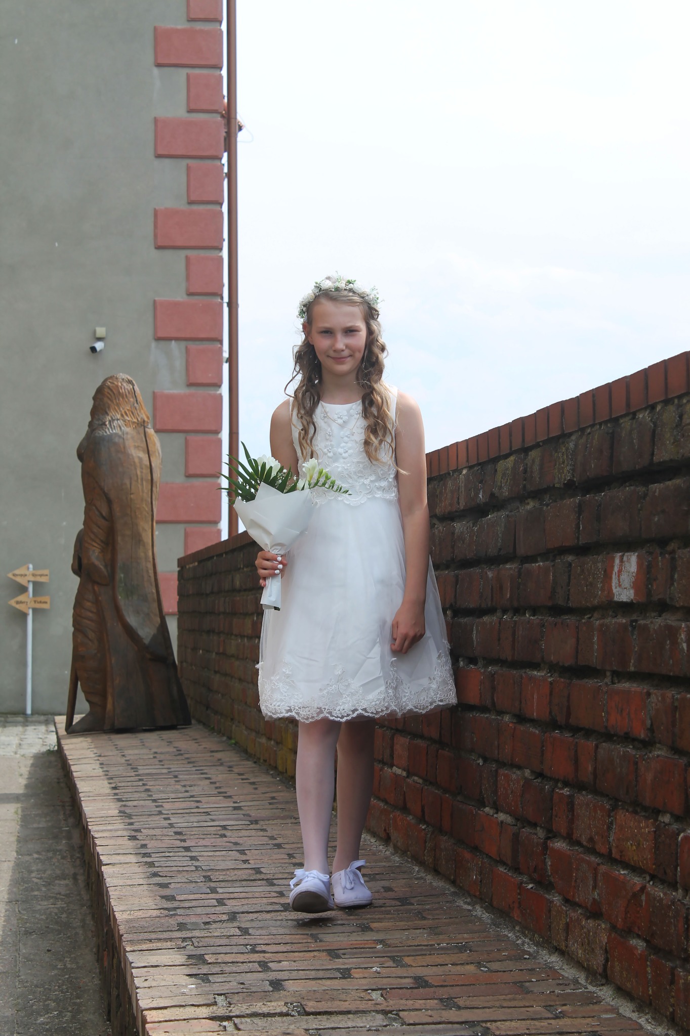 Dziewczynka w białej sukience komunijnej z wiankiem na głowie trzyma bukiet białych kwiatów, stojąc na ceglanym murku obok drewnianej rzeźby.