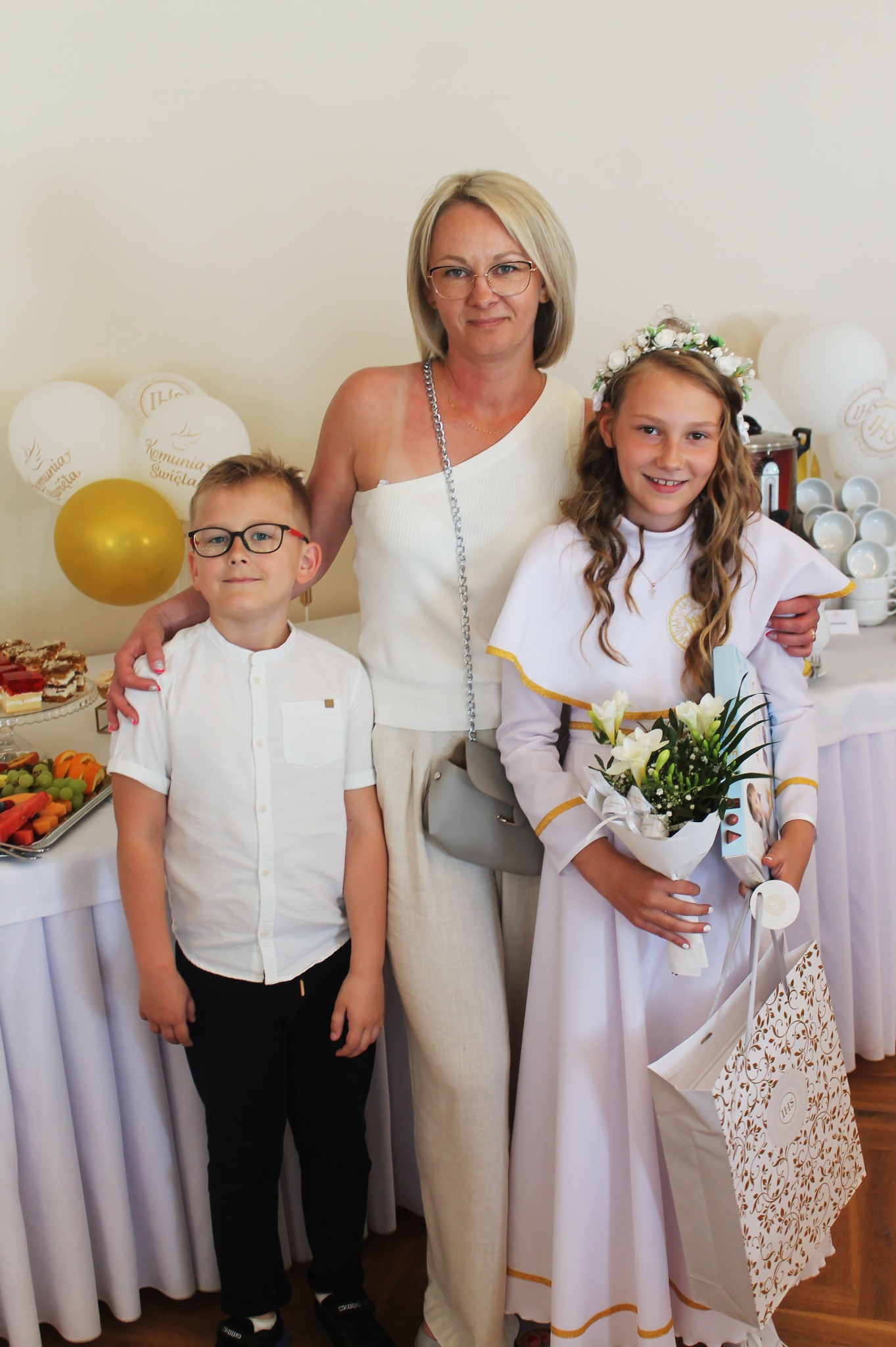 Dziewczynka w białej albie z wiankiem i bukietem lilii, pozuje z mamą i bratem na tle dekoracji komunijnych i stołu z przekąskami.