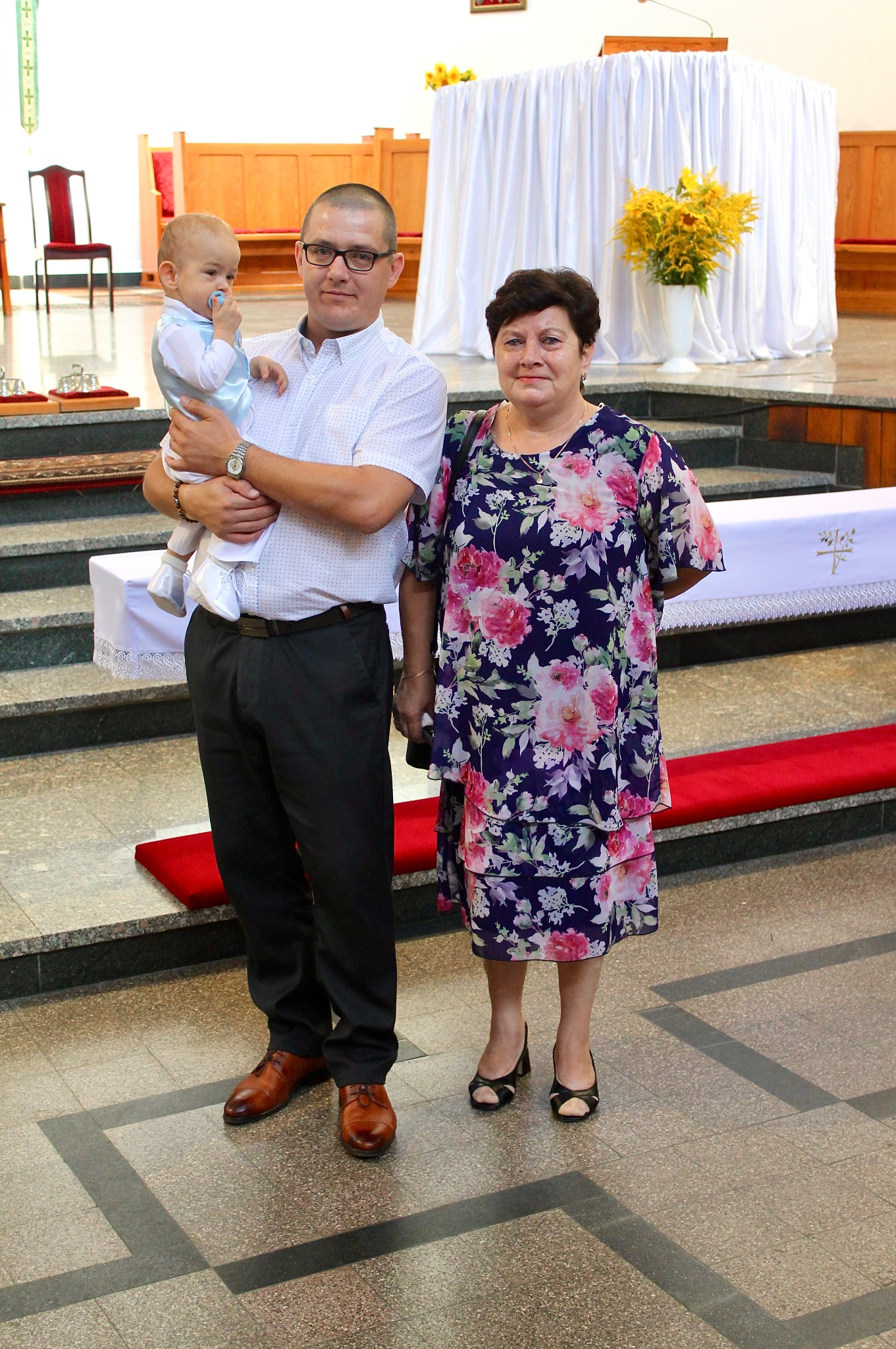 Rodzinne zdjęcie w kościele: mężczyzna trzyma niemowlę w jasnym ubranku, obok stoi kobieta w kwiecistej sukience. W tle ołtarz z białym obrusem i kwiatami.