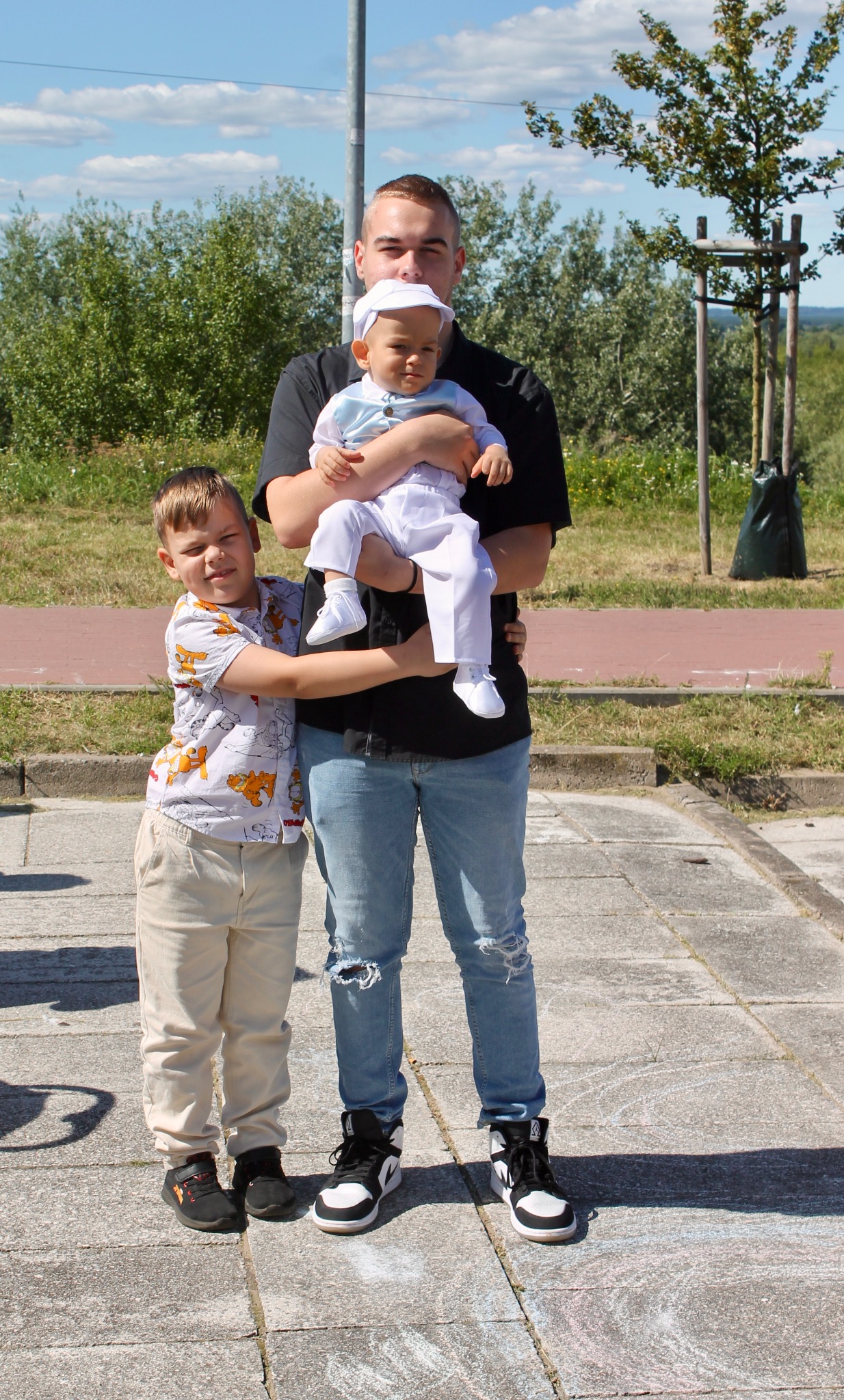 Mężczyzna w jeansach trzyma na rękach ubranego na biało niemowlaka. Obok stoi chłopiec w koszuli w lwy. W tle zieleń i błękitne niebo.