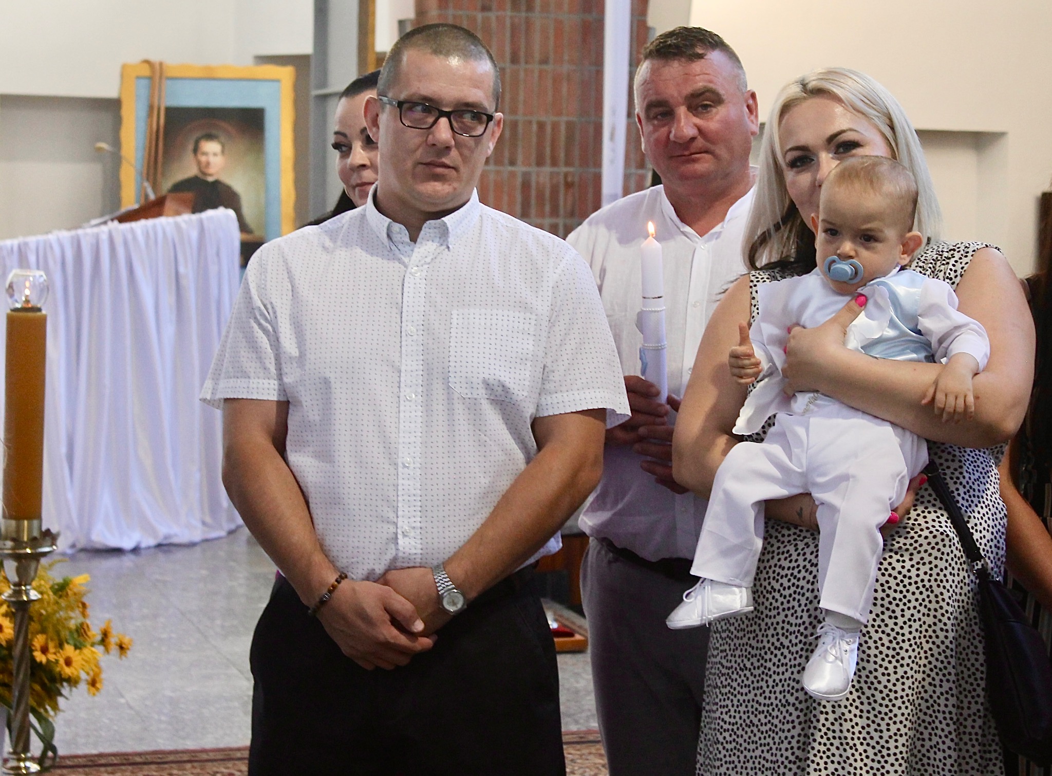 Rodzina z dzieckiem podczas uroczystości w kościele, w tle obraz religijny i świece. Mama trzyma niemowlę ubrane w biały strój.