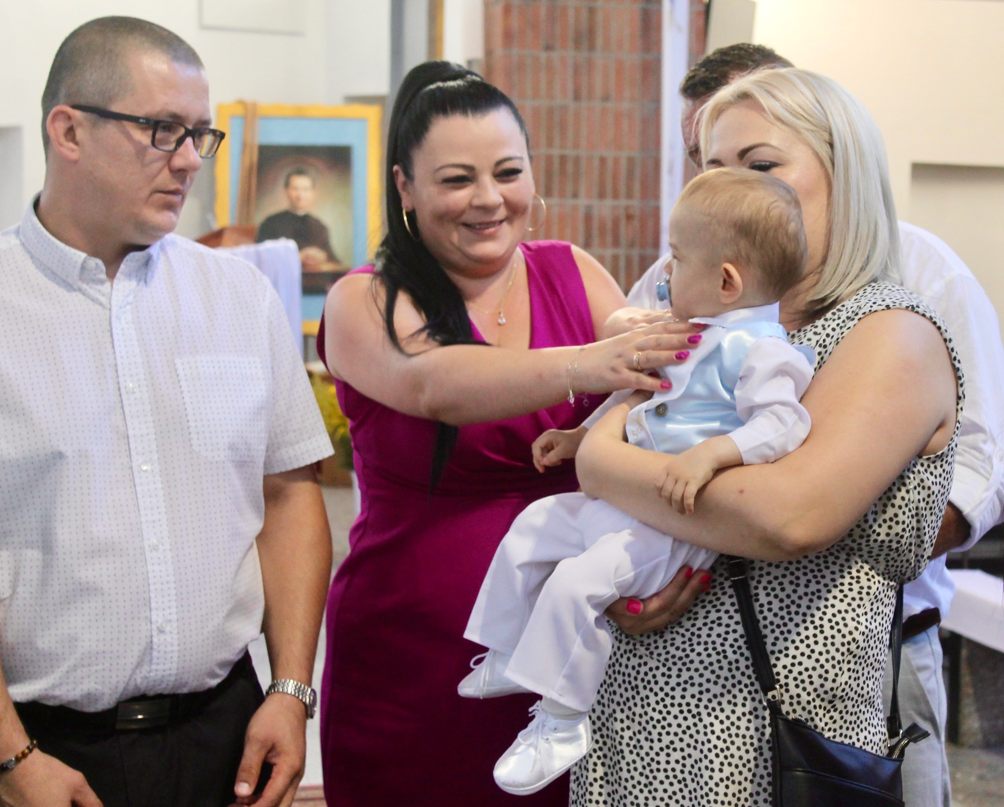 Uroczystość chrztu: kobieta trzyma niemowlę w błękitnej kamizelce, uśmiechnięta kobieta obok, mężczyzna z boku, w tle obraz w złotej ramie i ceglana ściana.