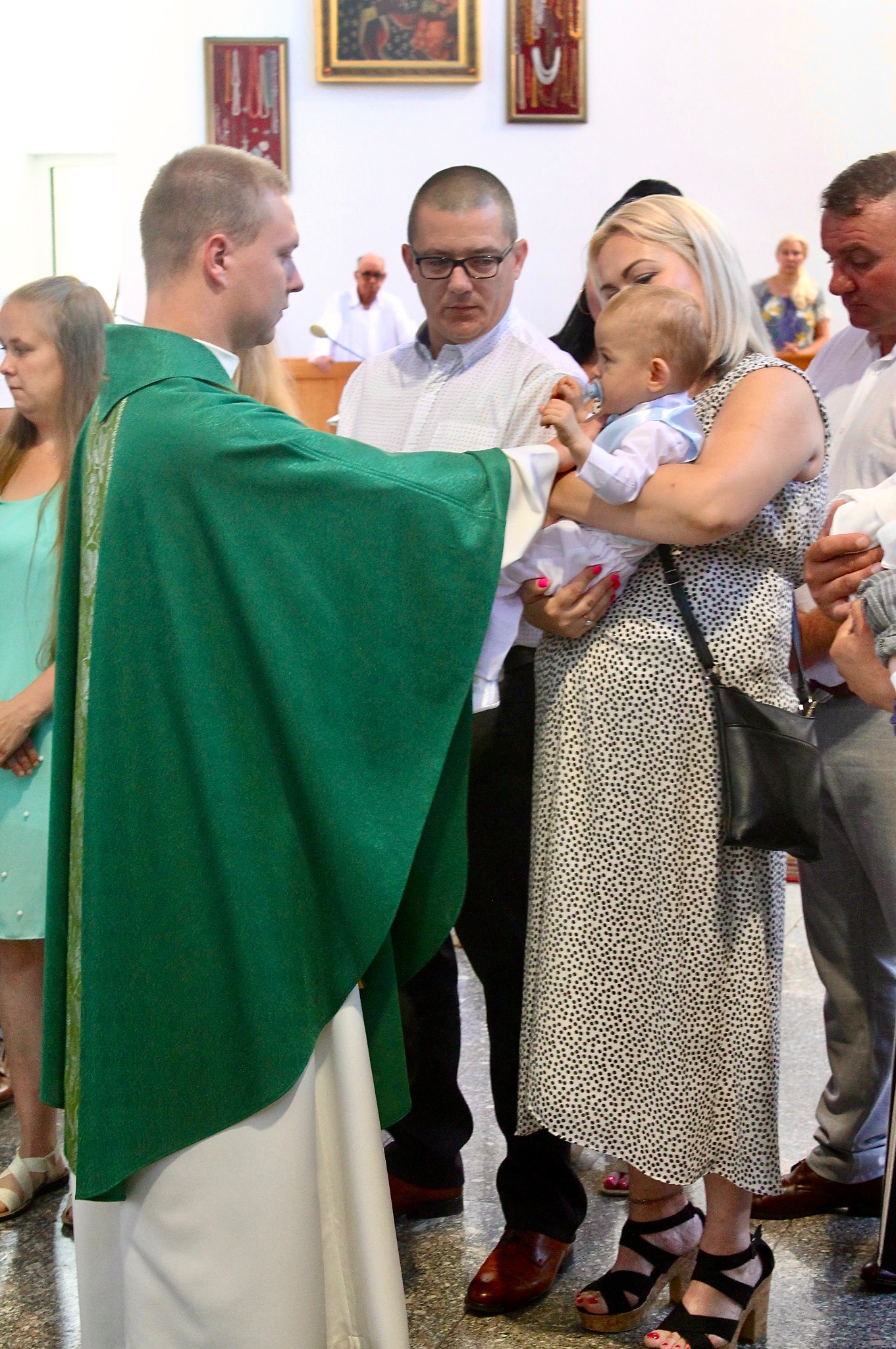 Ksiądz w zielonej szacie błogosławi niemowlę na rękach kobiety w sukience w kropki, w otoczeniu rodziny podczas uroczystości w kościele.