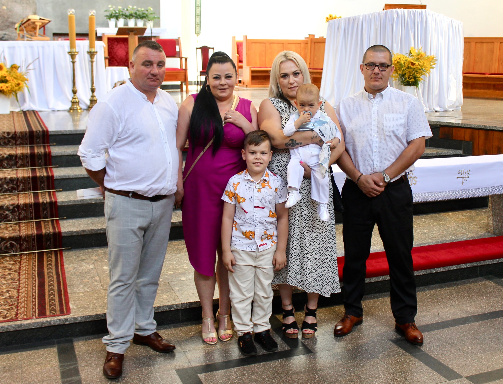 Rodzinne zdjęcie w kościele: dwoje dorosłych, dwoje dzieci i niemowlę. Uroczystość religijna, wnętrze kościoła, dywan, ołtarz w tle. Jasne oświetlenie.