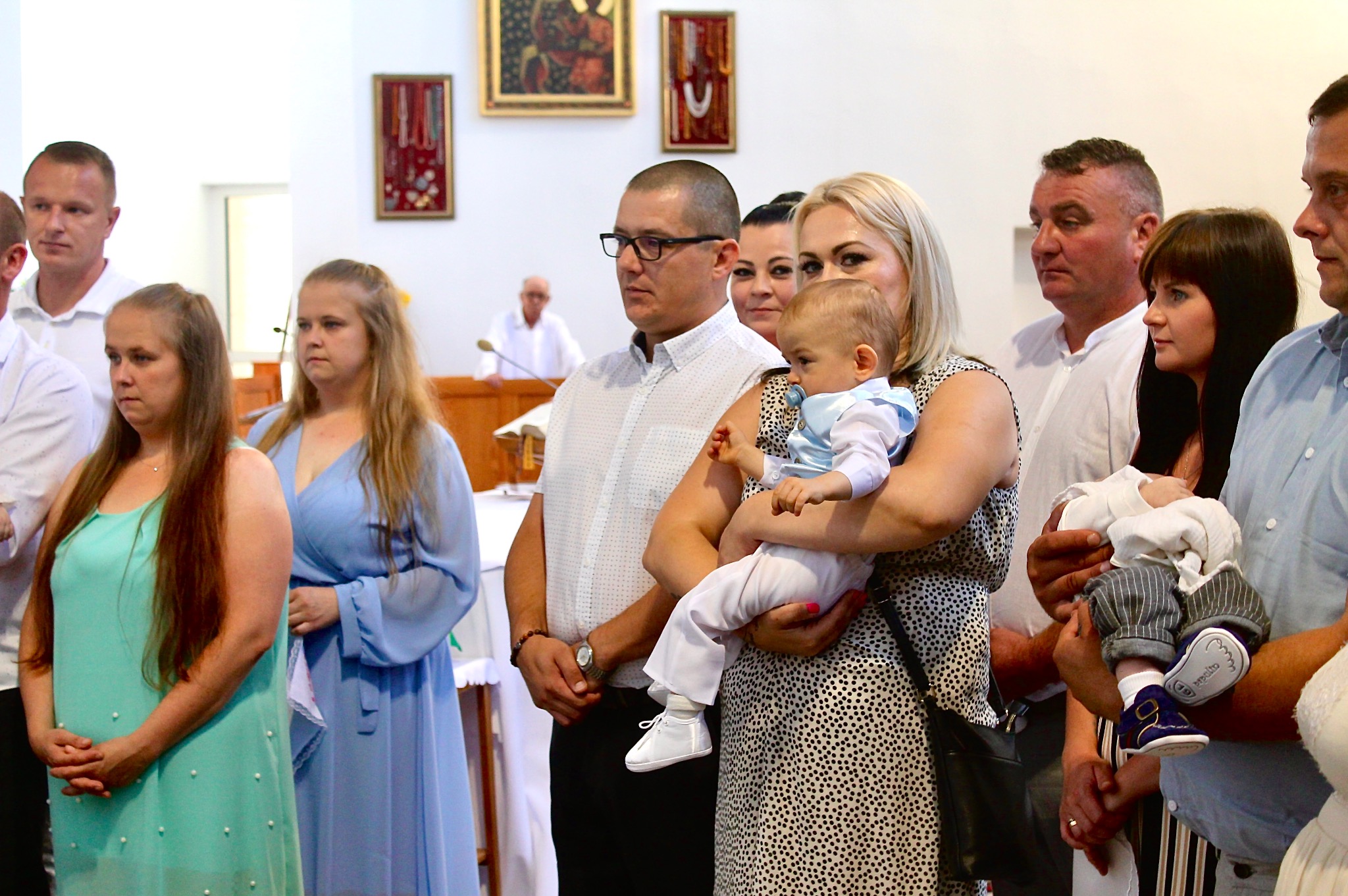 Goście podczas uroczystości w kościele, kobieta trzyma na rękach małe dziecko ubrane w biały garnitur. W tle obrazy religijne.