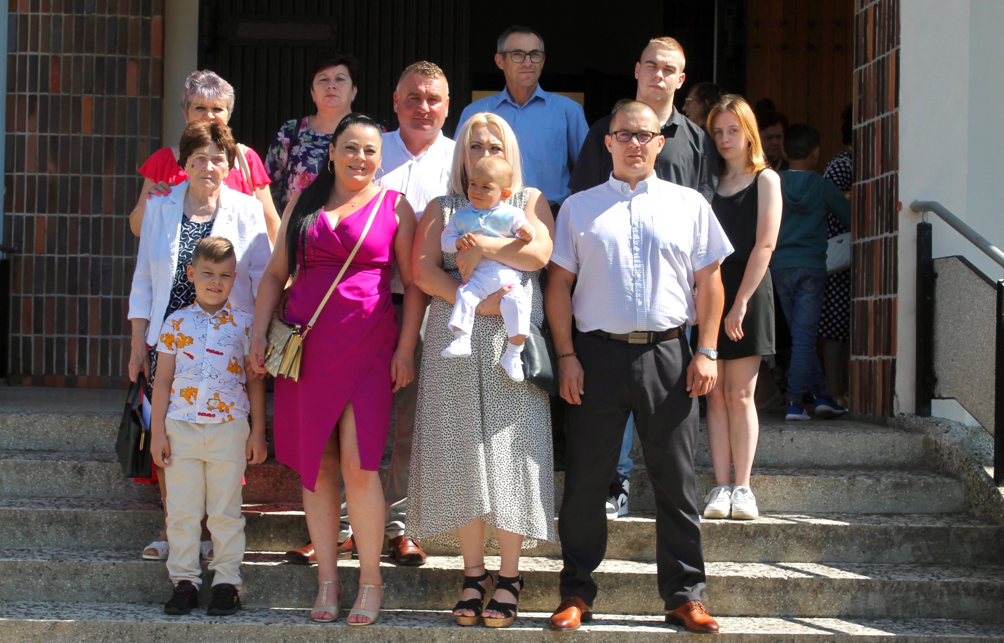 Grupa ludzi w różnym wieku, ubranych elegancko, pozuje na schodach przed budynkiem. Widoczna kobieta trzymająca małe dziecko. Uroczystość rodzinna.