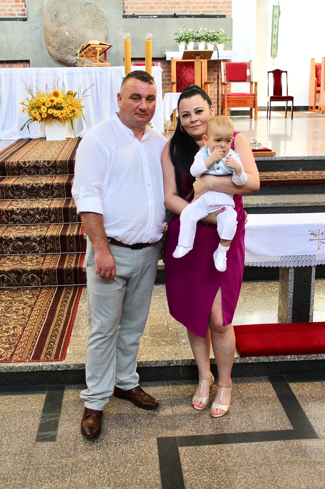 Rodzinne zdjęcie w kościele po uroczystości chrztu, rodzice z dzieckiem na tle ołtarza i dekoracji kwiatowych. Ujęcie z bliska, naturalne światło.