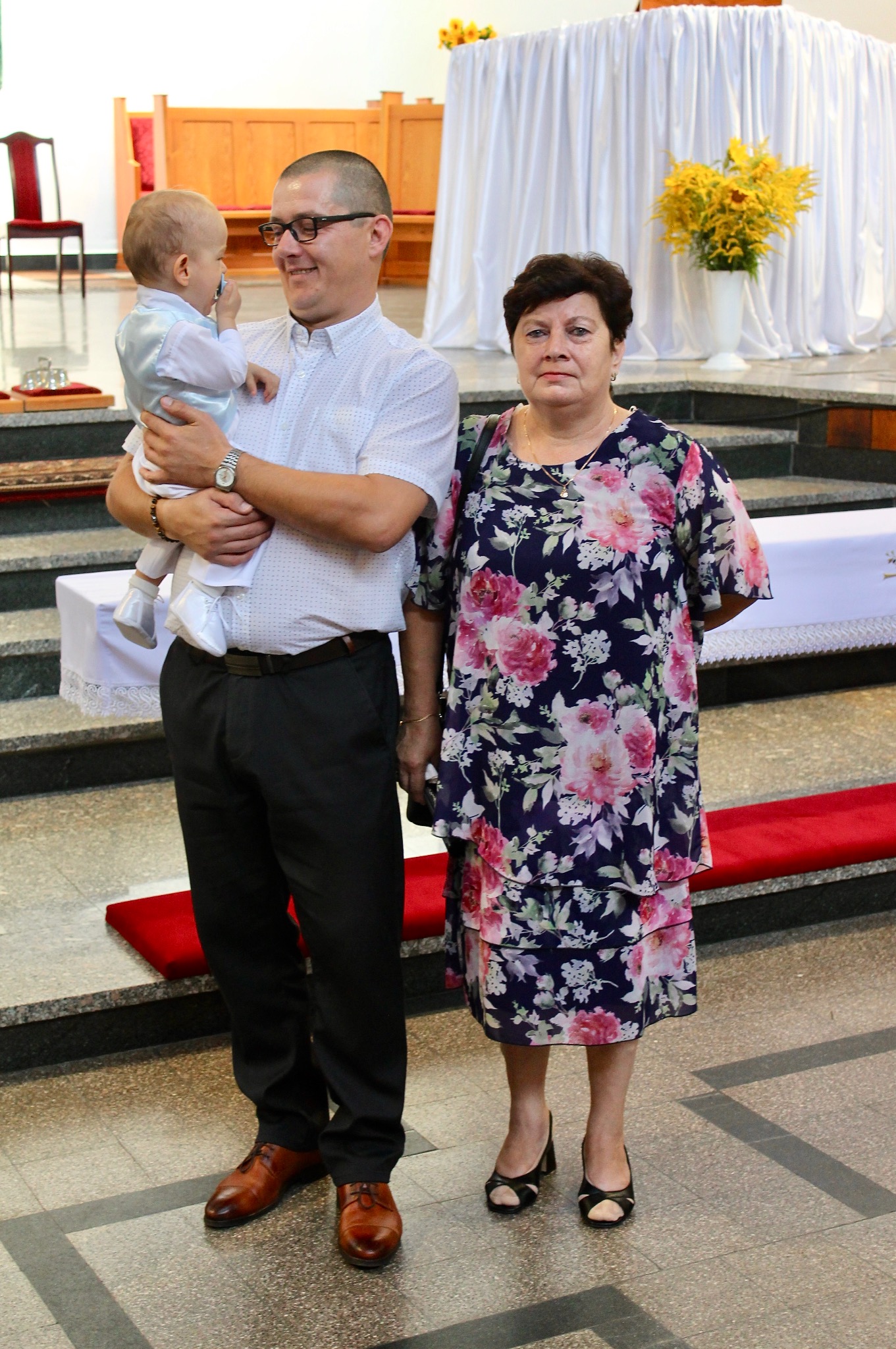 Uroczystość w kościele: mężczyzna z dzieckiem na rękach i kobieta w kwiecistej sukience stoją na tle ołtarza z białymi zasłonami i żółtymi kwiatami.