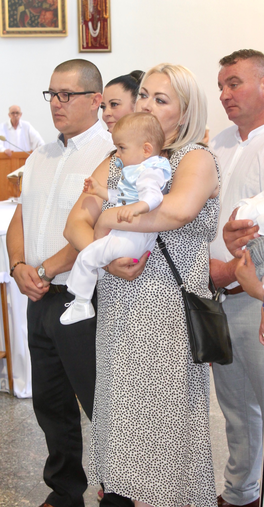 Rodzina podczas uroczystości kościelnej, matka trzyma niemowlę w błękitnym ubranku, obok stoi mężczyzna w okularach, w tle widoczny ksiądz.