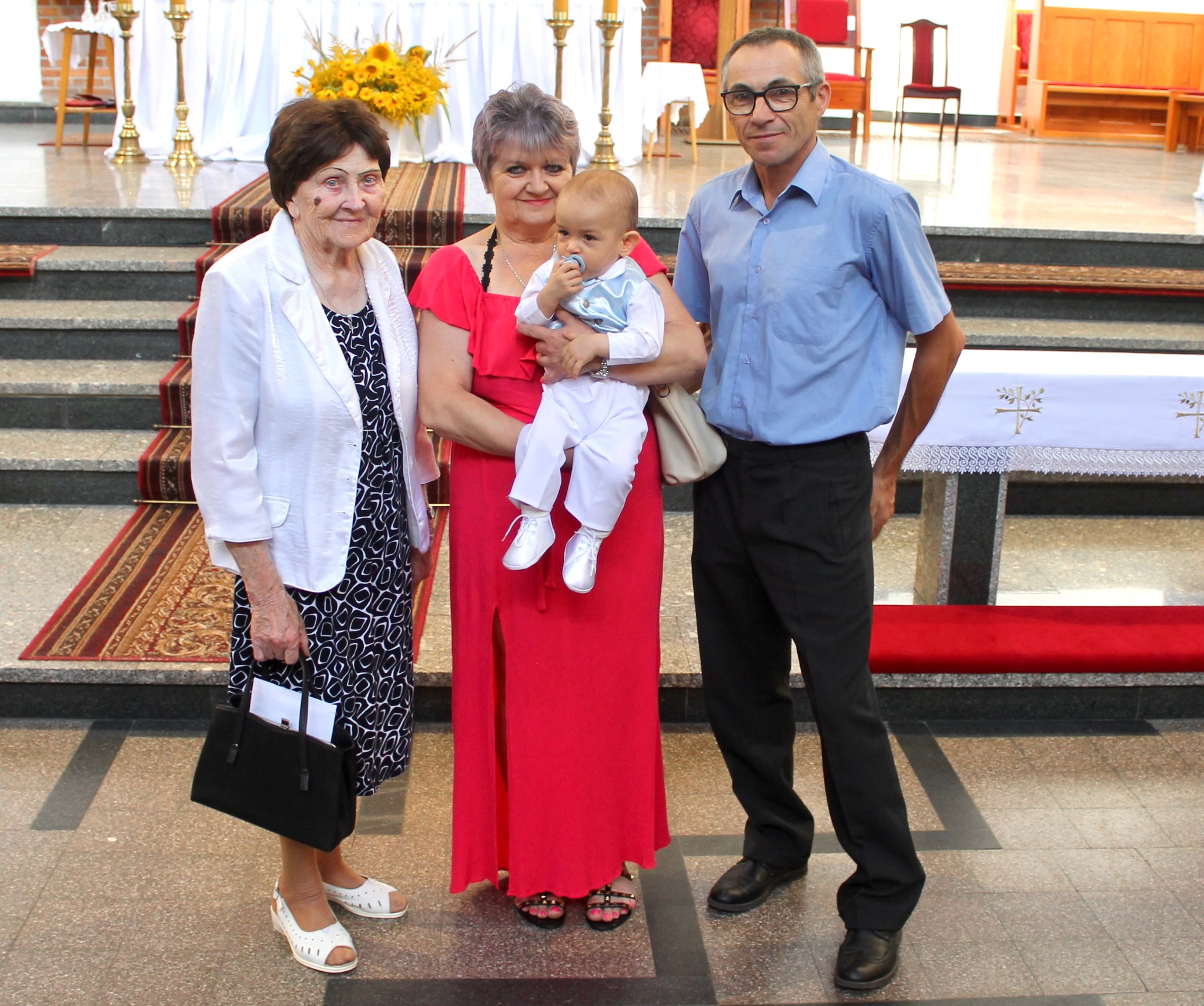 Rodzinne zdjęcie w kościele: babcia z torebką, mama w czerwonej sukience trzymająca niemowlę w błękitnej kamizelce i tata w niebieskiej koszuli. Uroczystość rodzinna.