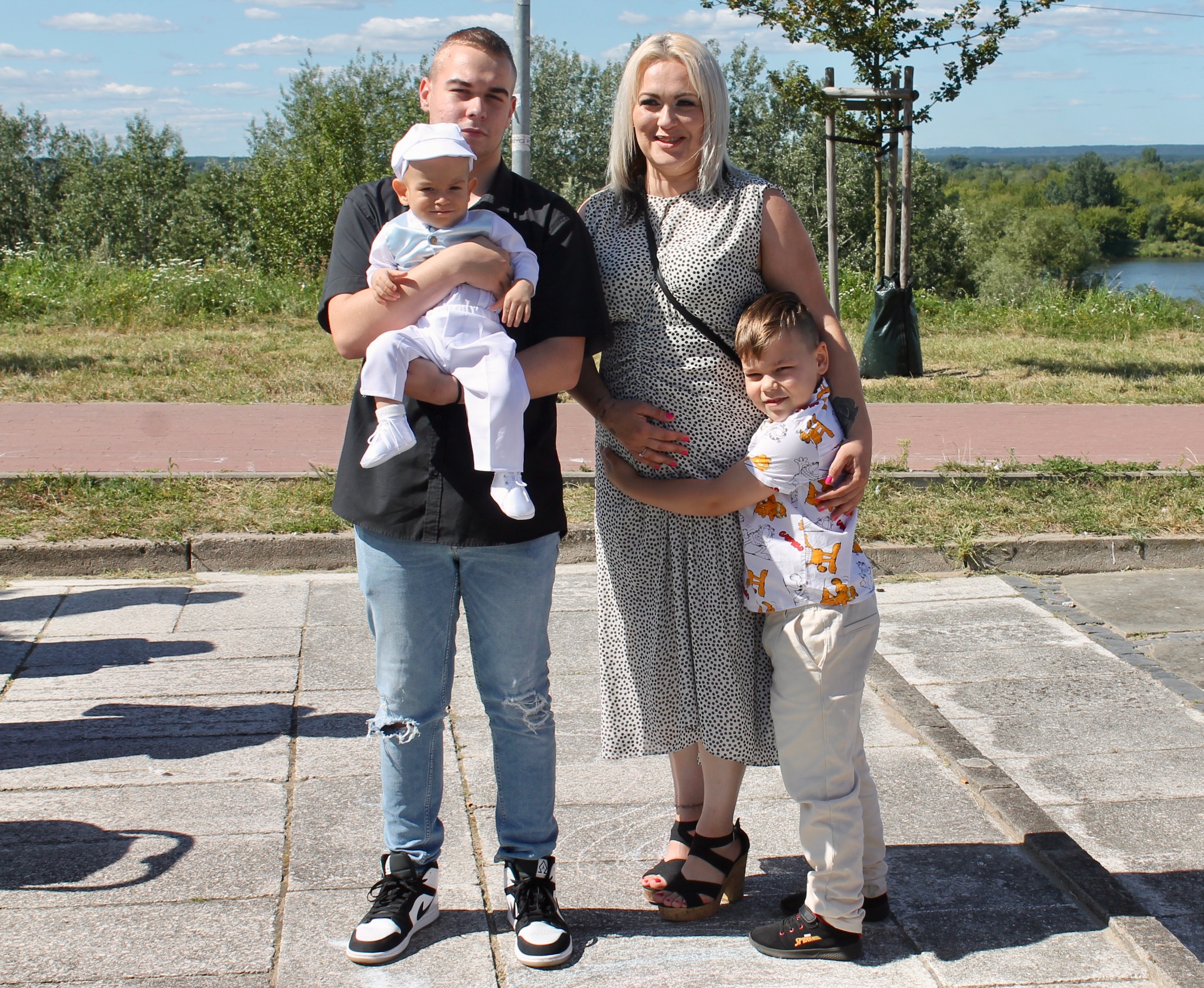 Rodzinne zdjęcie plenerowe: młody mężczyzna trzyma niemowlę w białym ubranku, obok kobieta w sukience i starszy chłopiec. W tle zieleń i rzeka.