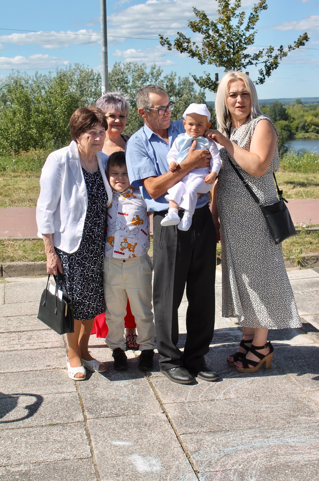 Rodzinne zdjęcie grupowe z dzieckiem w centrum, wykonane w plenerze w słoneczny dzień, z widokiem na rzekę w tle.