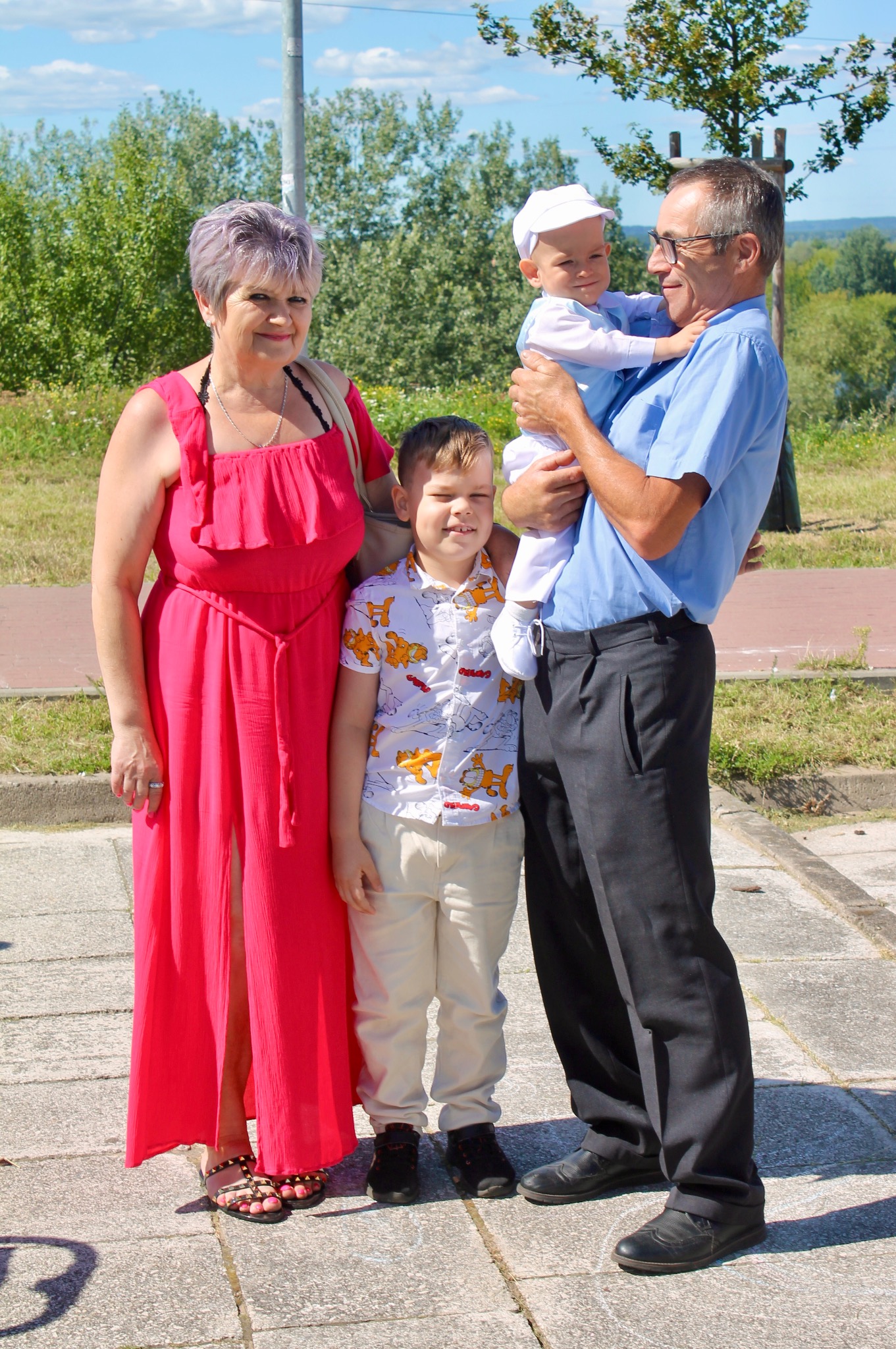 Rodzinne zdjęcie na zewnątrz: kobieta w czerwonej sukience, chłopiec w koszuli z motywem zwierzęcym, mężczyzna trzymający niemowlę w białym ubranku i czapeczce.