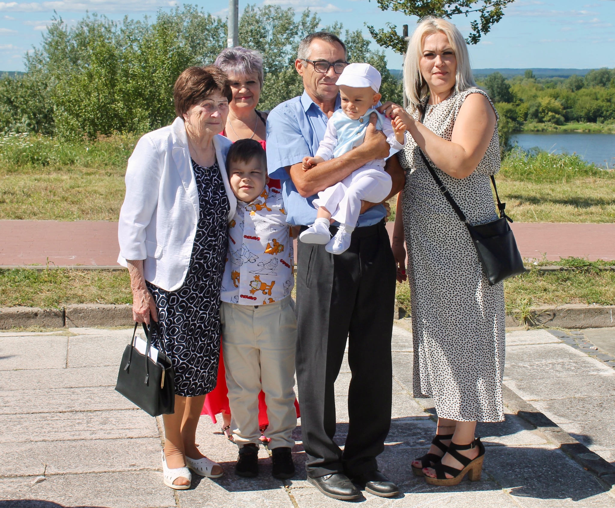 Rodzinne zdjęcie grupowe z dzieckiem w centrum, na tle zieleni i rzeki. Ubrania eleganckie, okazjonalne. Dziecko w białym ubranku i czapeczce.