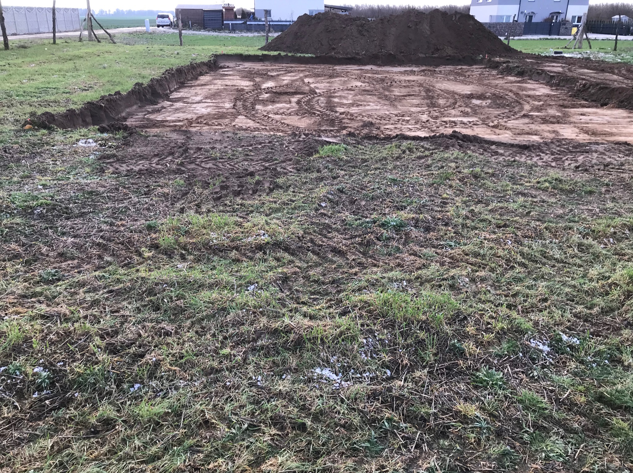 Przygotowany teren pod budowę, widoczne ślady opon, wykopana prostokątna przestrzeń z pryzmą ziemi w tle, trawa z resztkami śniegu na pierwszym planie.