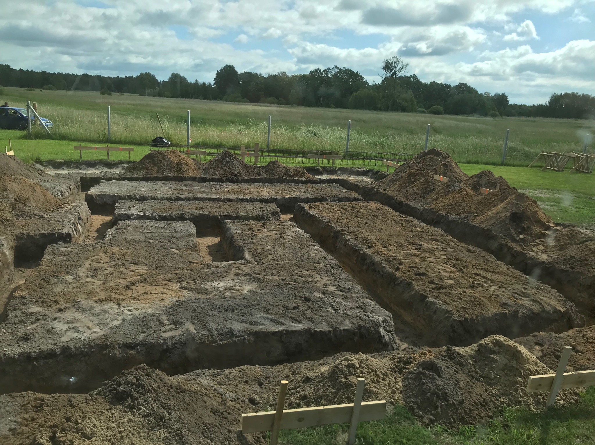 Przygotowany teren pod budowę: wykopane rowy pod fundamenty w ziemi, z hałdami ziemi obok, widoczne drewniane elementy szalunku i zbrojenie, w tle łąka i las.