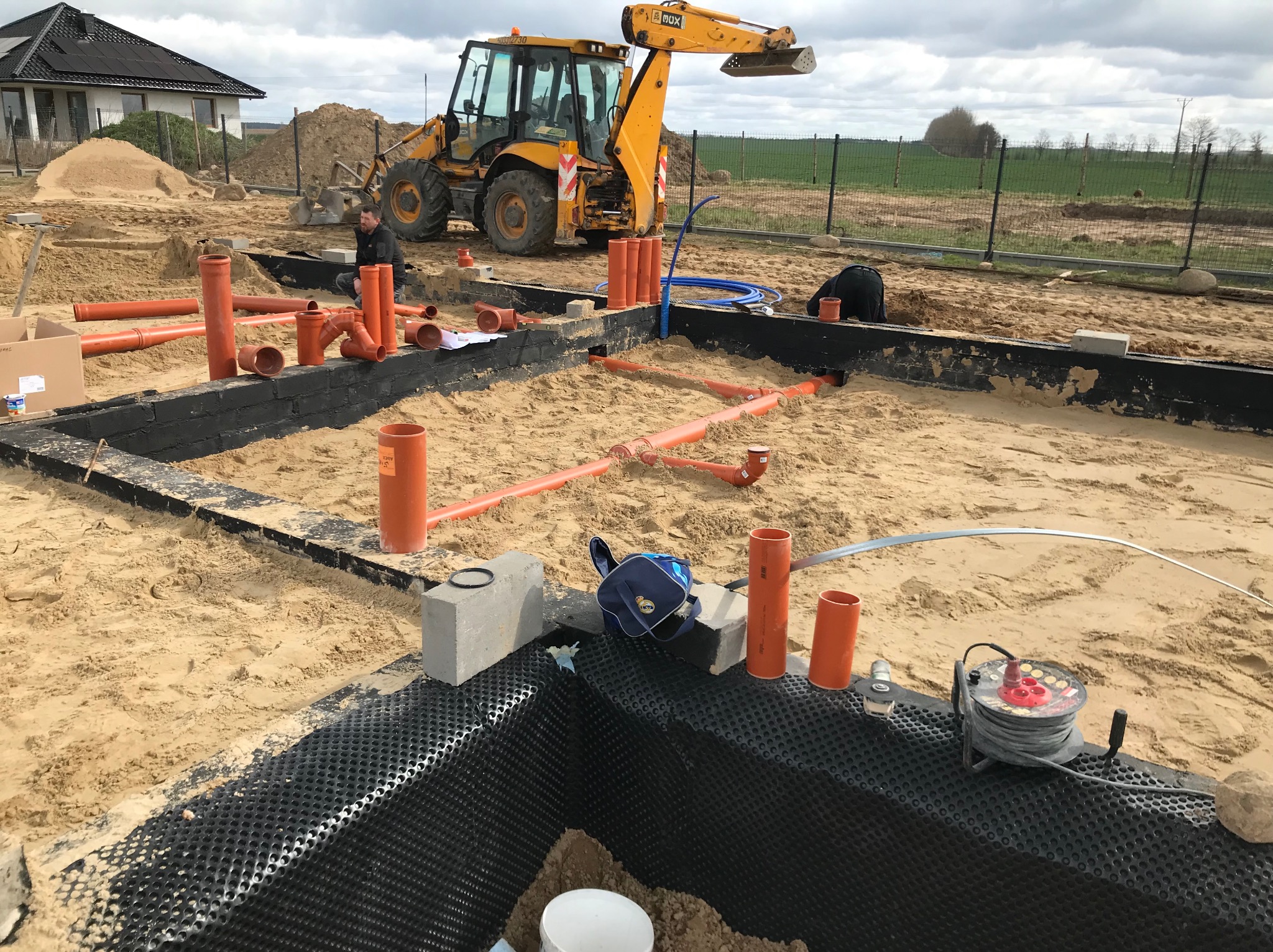 Żółta koparko-ładowarka na placu budowy, widoczne rury kanalizacyjne, izolacja fundamentów i pracownicy w trakcie montażu instalacji w wykopie.