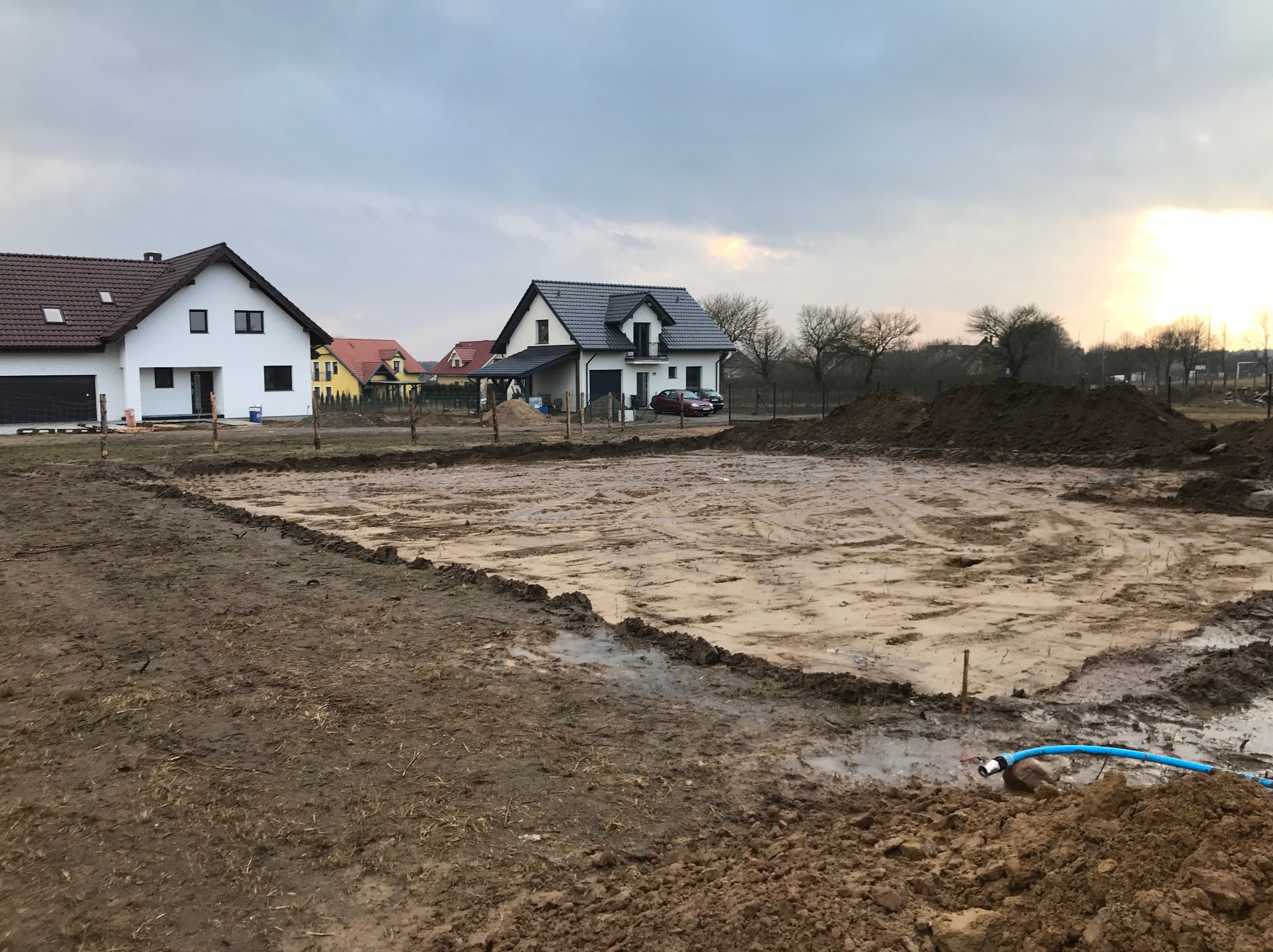 Przygotowany teren pod budowę domu, widoczne wytyczone granice fundamentów, świeżo rozkopana ziemia i wąż doprowadzający wodę, w tle zabudowa jednorodzinna.