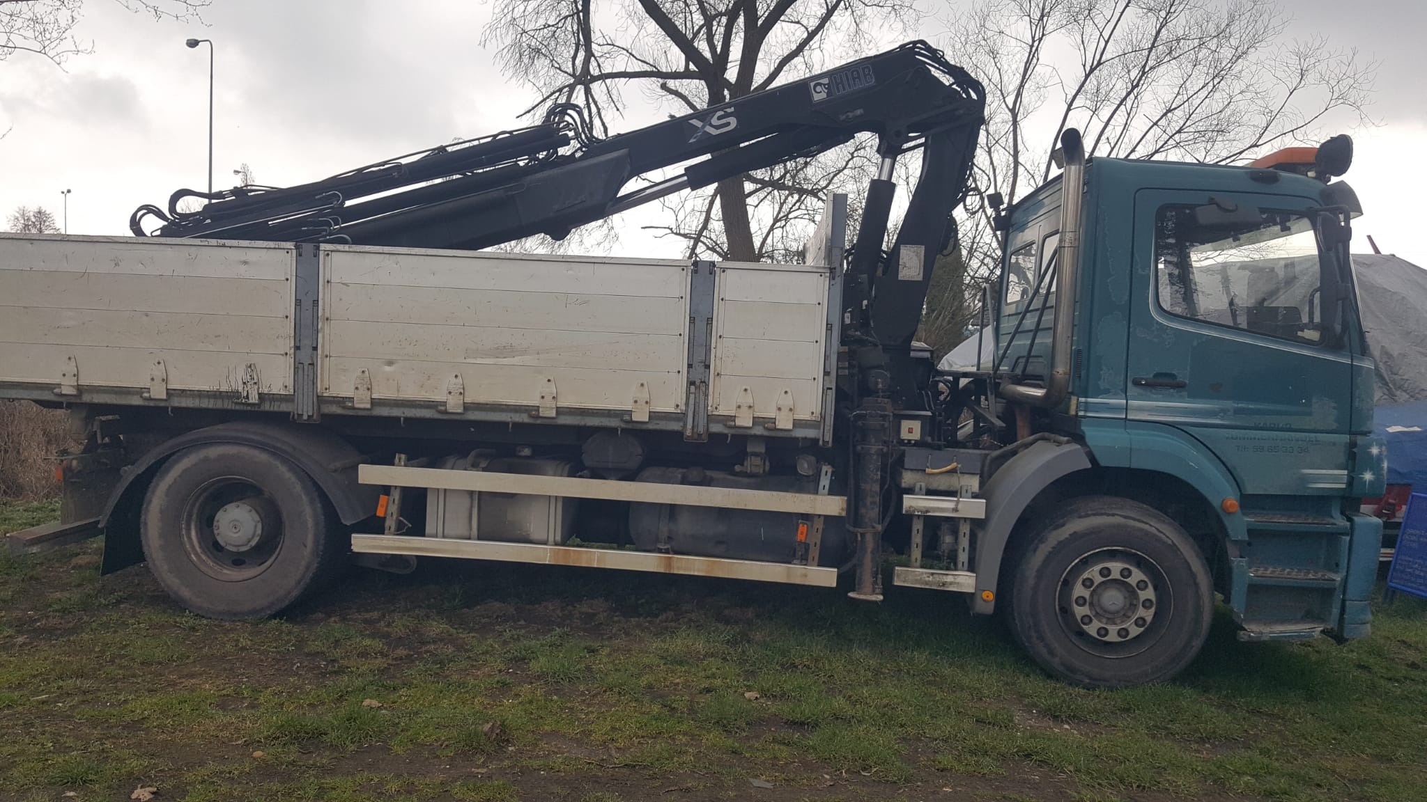 Ciężarówka z dźwigiem HIAB XS, zaparkowana na trawiastym terenie, widoczne drzewa bez liści w tle.