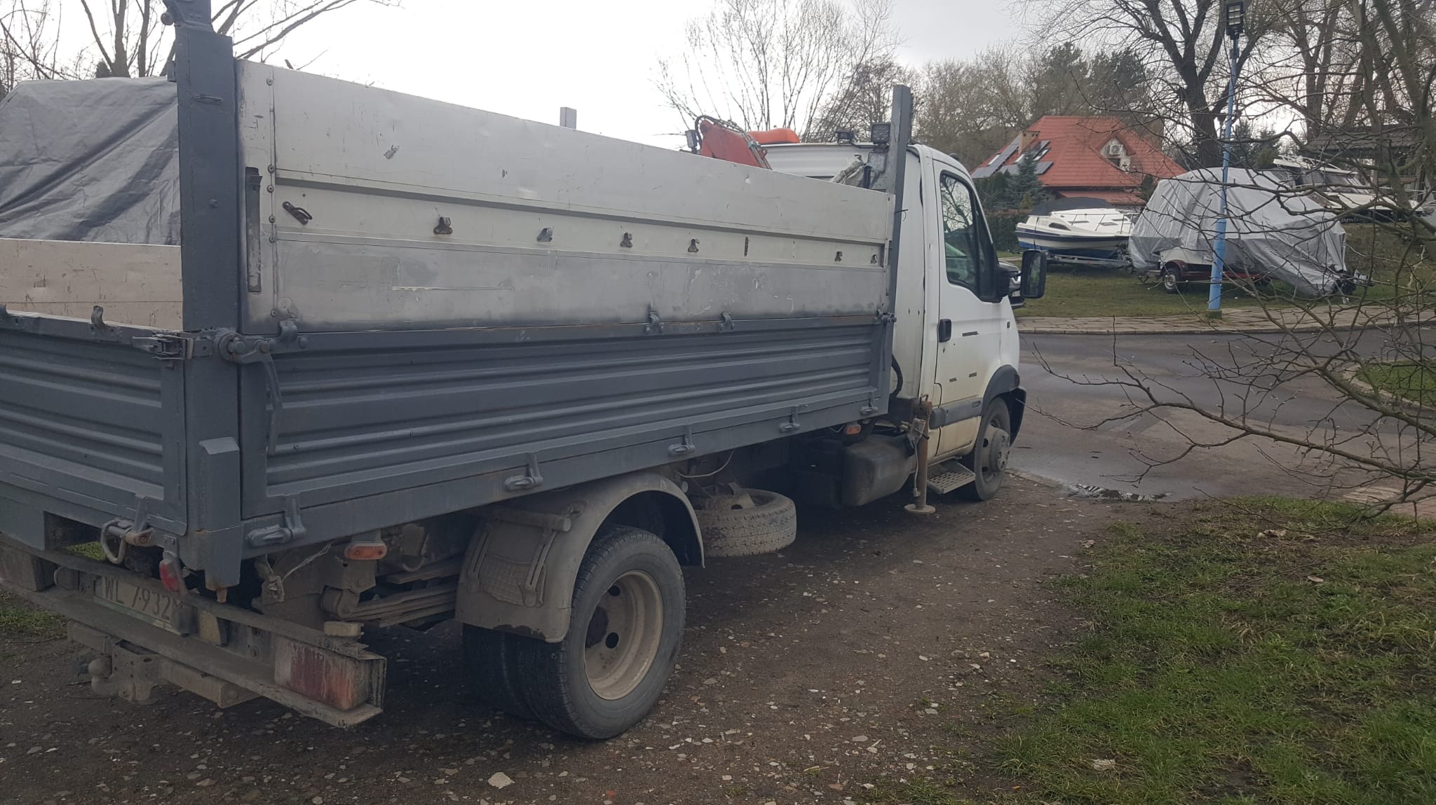 Ciężarówka typu wywrotka z plandeką na pace, zaparkowana na ziemnej nawierzchni, w tle domy i przykryte plandekami łodzie.