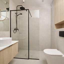 Wito - Remonty - Nowoczesna łazienka z kabiną prysznicową typu walk-in z czarnymi profilami, umywalką nablatową na szafce z drewnopodobnym fornirem oraz wiszącą toaletą z czarnym przyciskiem spłukującym.