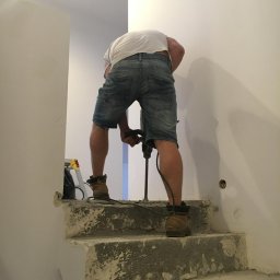 Wito - Remonty - Mężczyzna w dżinsowych szortach wierci w ścianie na schodach, widoczny stary beton i przygotowania do remontu.