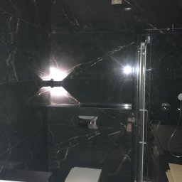 Wito - Remonty - Nowoczesna łazienka z czarnymi płytkami imitującymi marmur, podświetlana ściana, wisząca toaleta i szklana kabina prysznicowa.